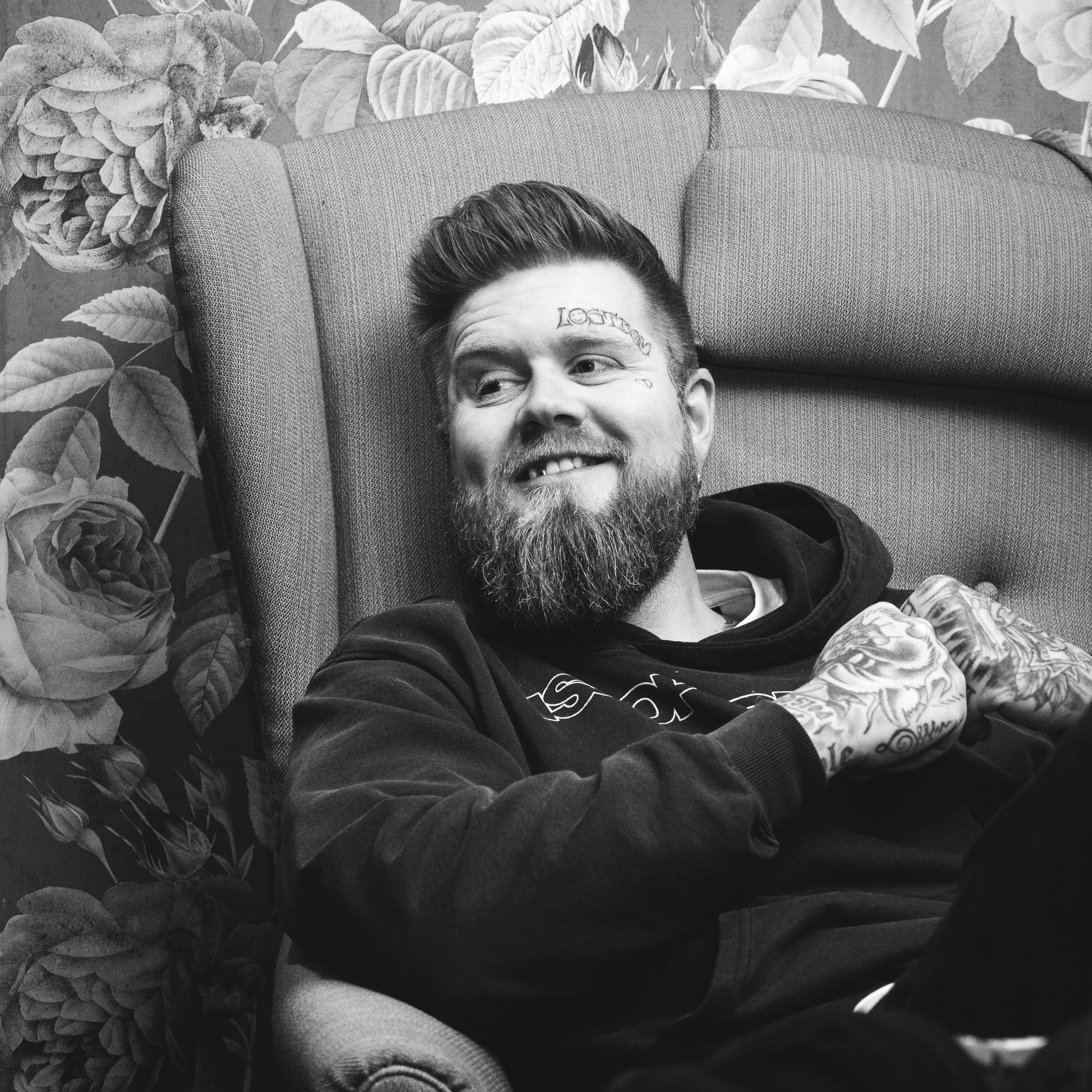 En mann med skjegg og tatoveringer ligger i en lenestol med en blomsterdekket vegg i bakgrunnen.