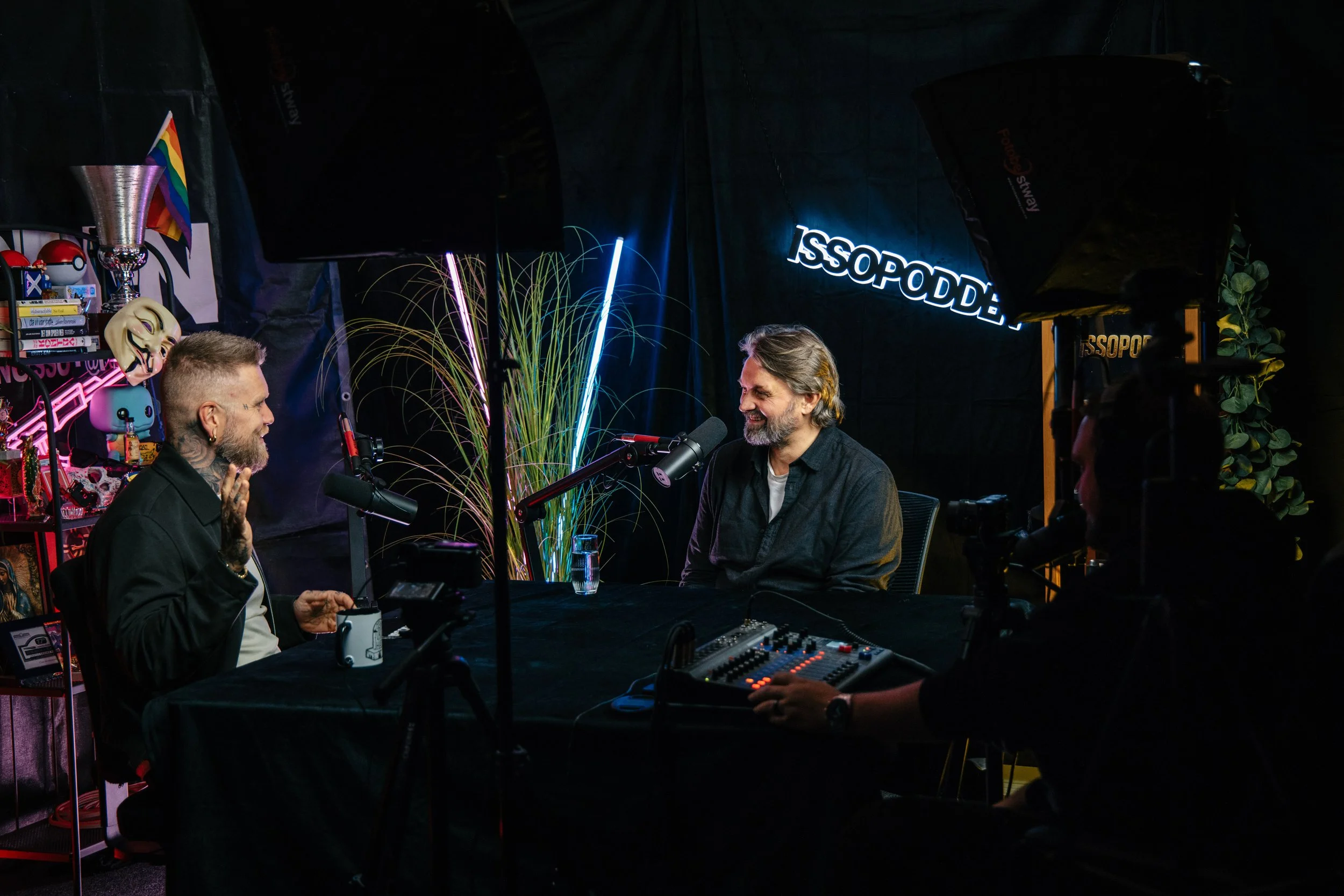 To personer snakker i en podkastepisode, sittende ved et svart bord med mikrofoner, omgitt av ulike dekorasjoner og belysning. En person har skjegg og grått hår, den andre har tatoveringer og kort hår.
