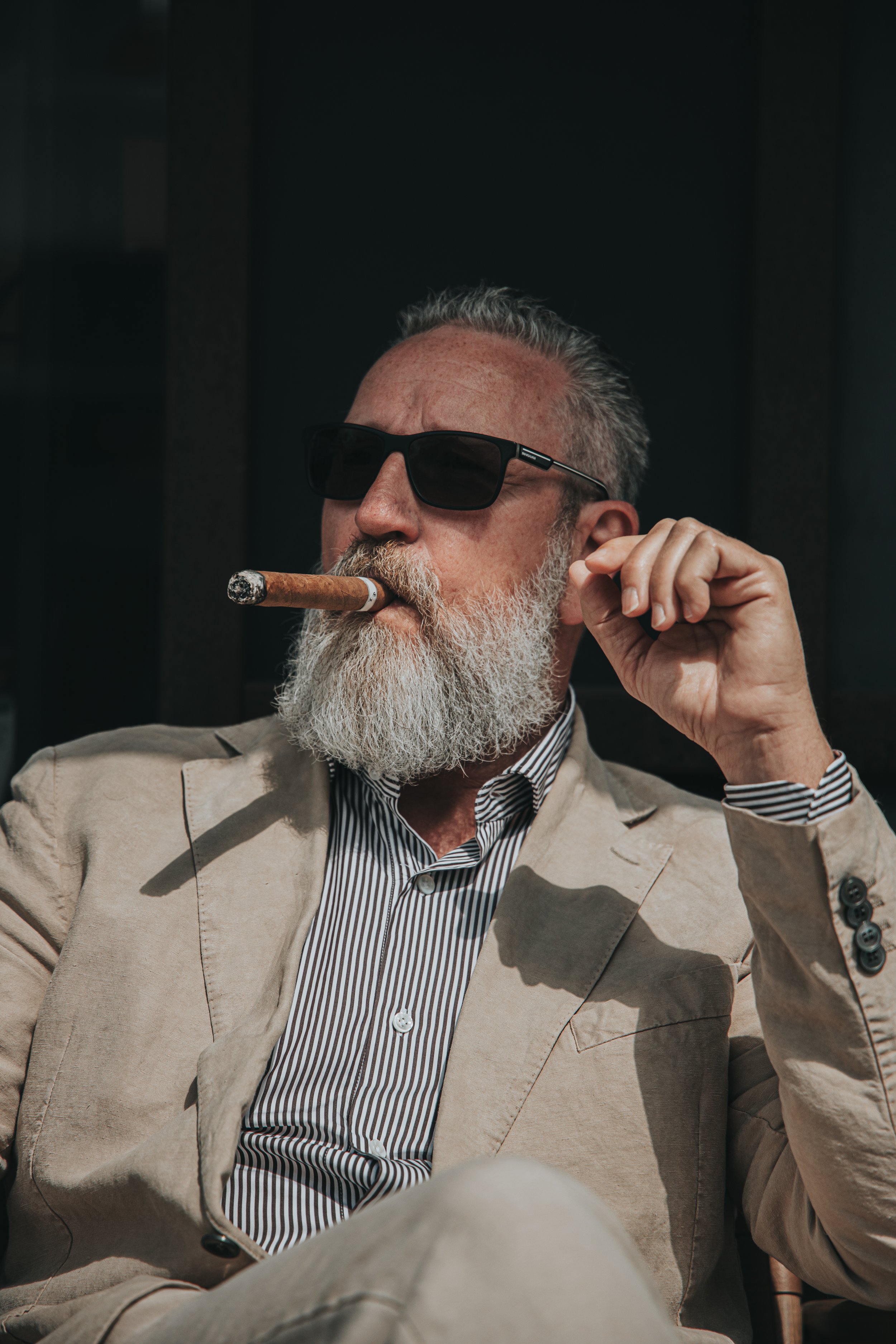 En eldre mann med grått skjegg, solbriller og en sigar i munnen, iført en beige dress og en stripet skjorte.