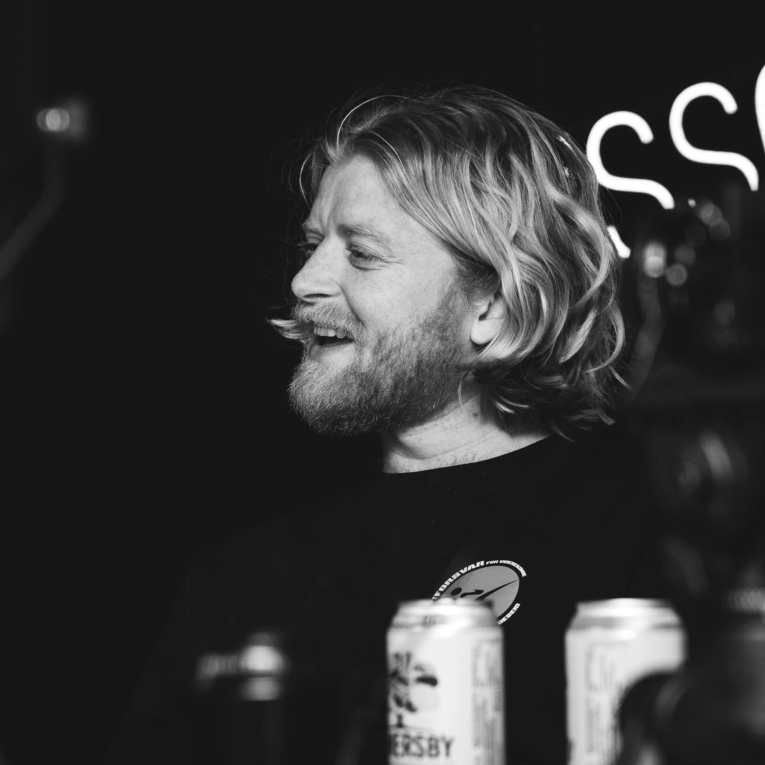 Et smilende mannsansikt i profil, med langt, bølget hår og skjegg, tatt i svart-hvitt foran en mørk bakgrunn med noe belysning og utstyr i forgrunnen.