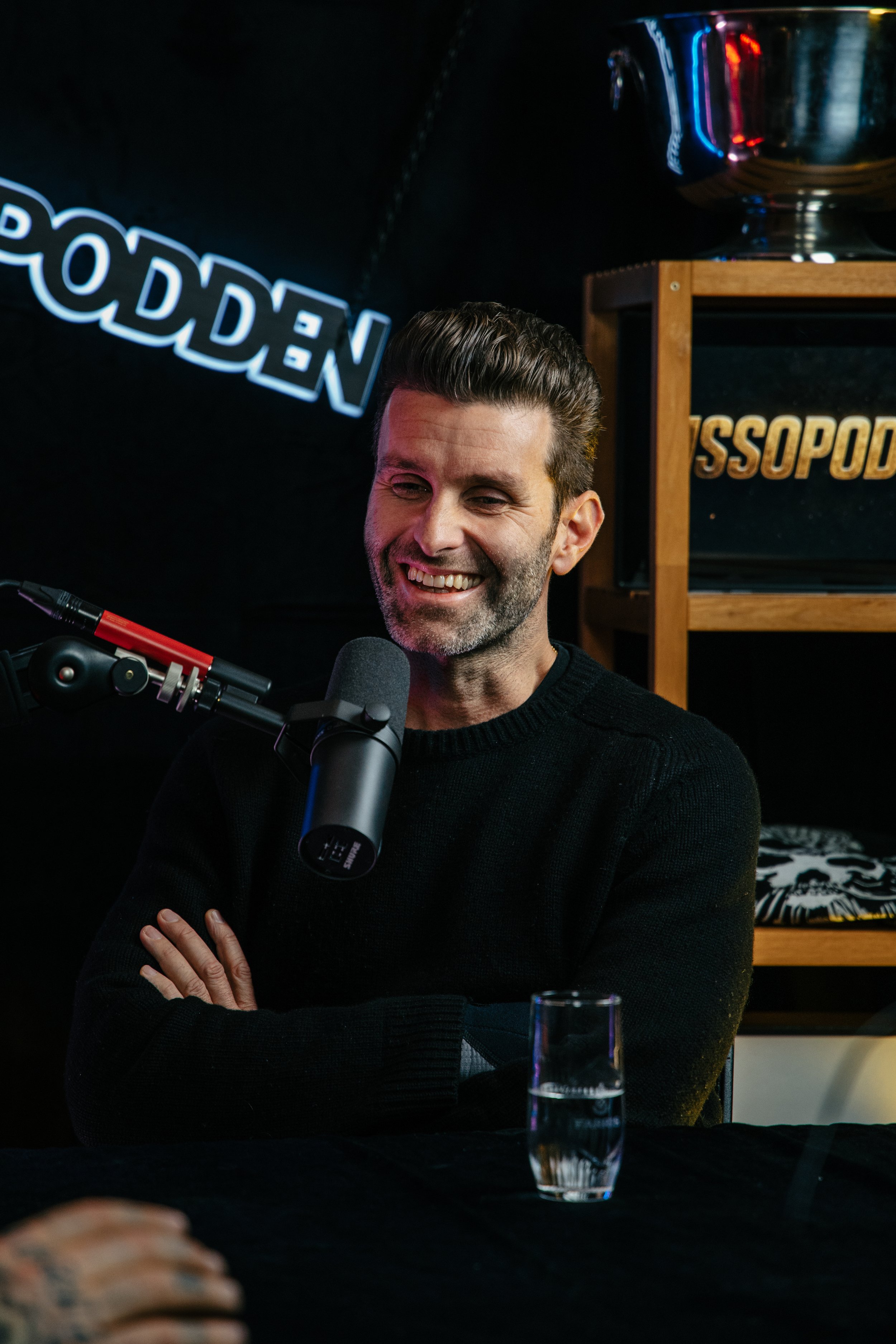 En mann smiler mens han snakker i en mikrofon under en innendørs innspilling eller podcast. Han har mørkt hår og skjegg, og ser ut til å være kledd i en svart genser. I bakgrunnen er det en hyller med et pokal og en tekst som sier "ROSSOP". Det er og