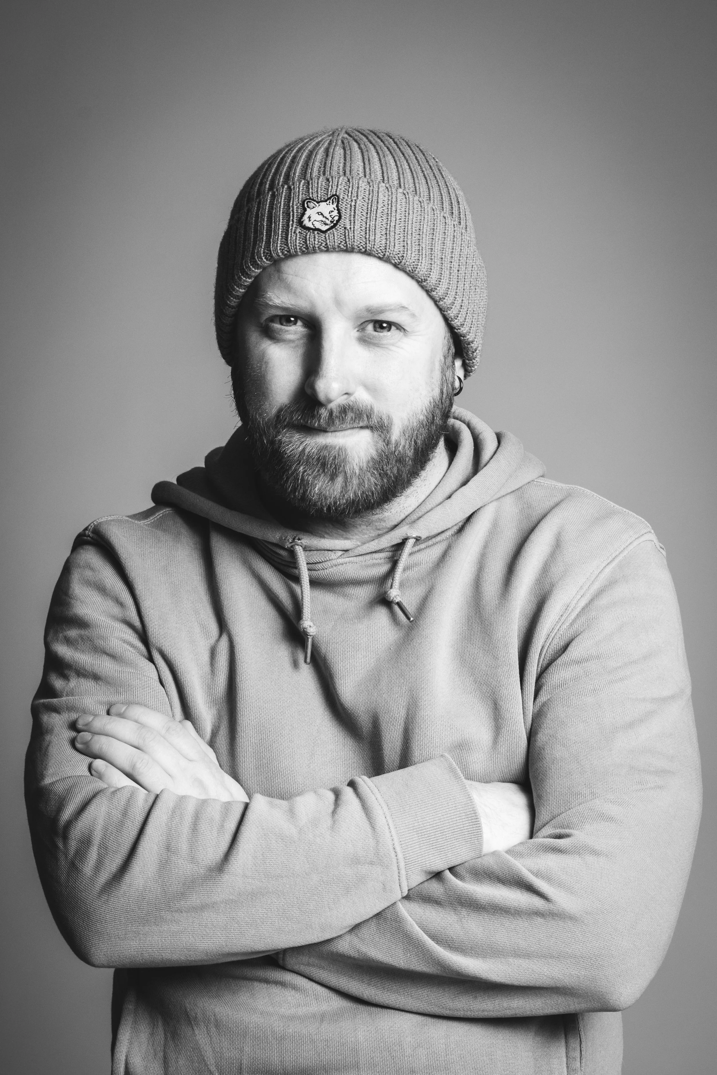Black and white portrait of a bearded man Espen Tønnesen wearing a knit beanie and hoodie, crossing his arms and looking at the camera.