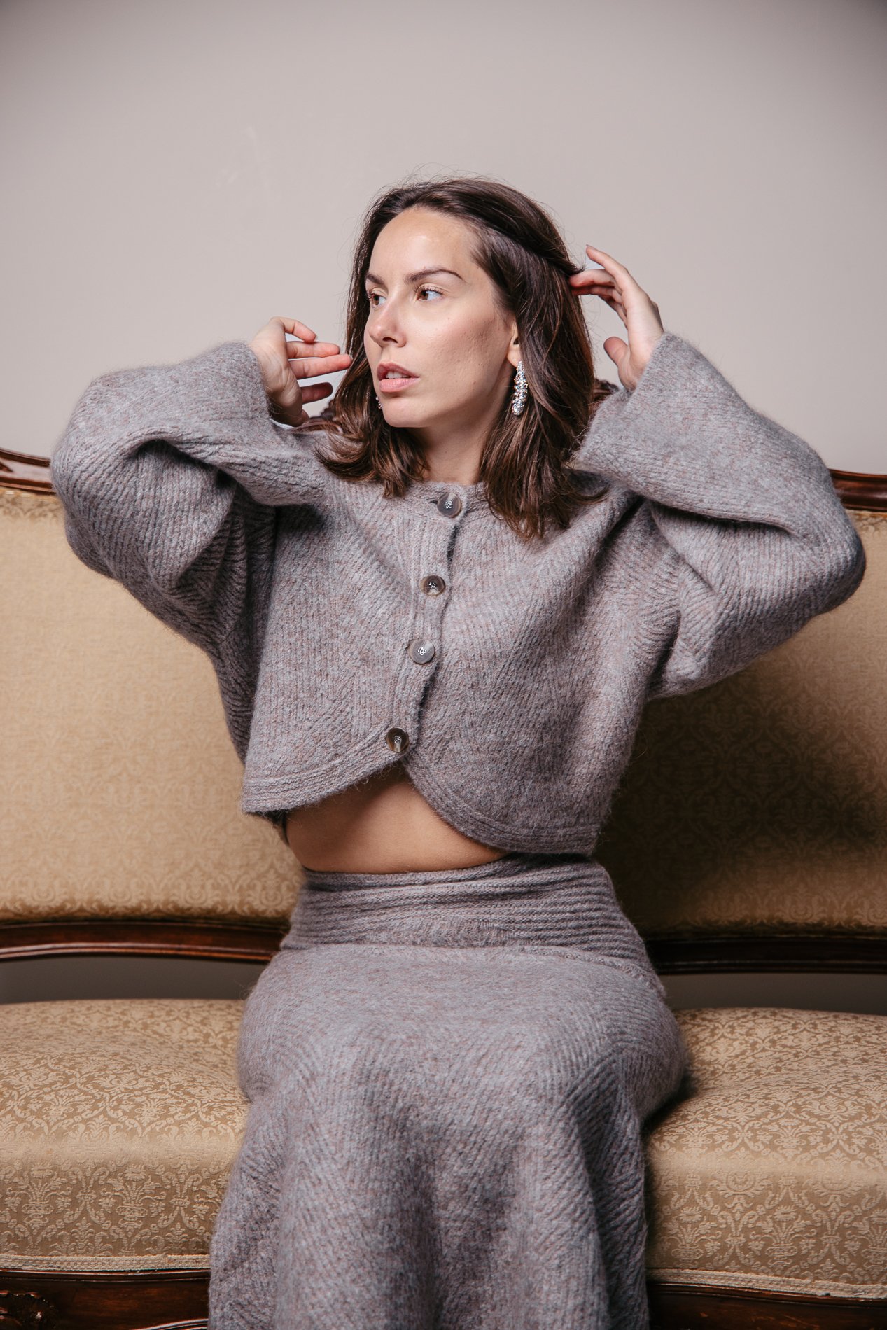 A woman with dark brown hair in a gray cardigan and matching skirt, sitting on a vintage sofa, adjusting her hair, wearing large earrings.