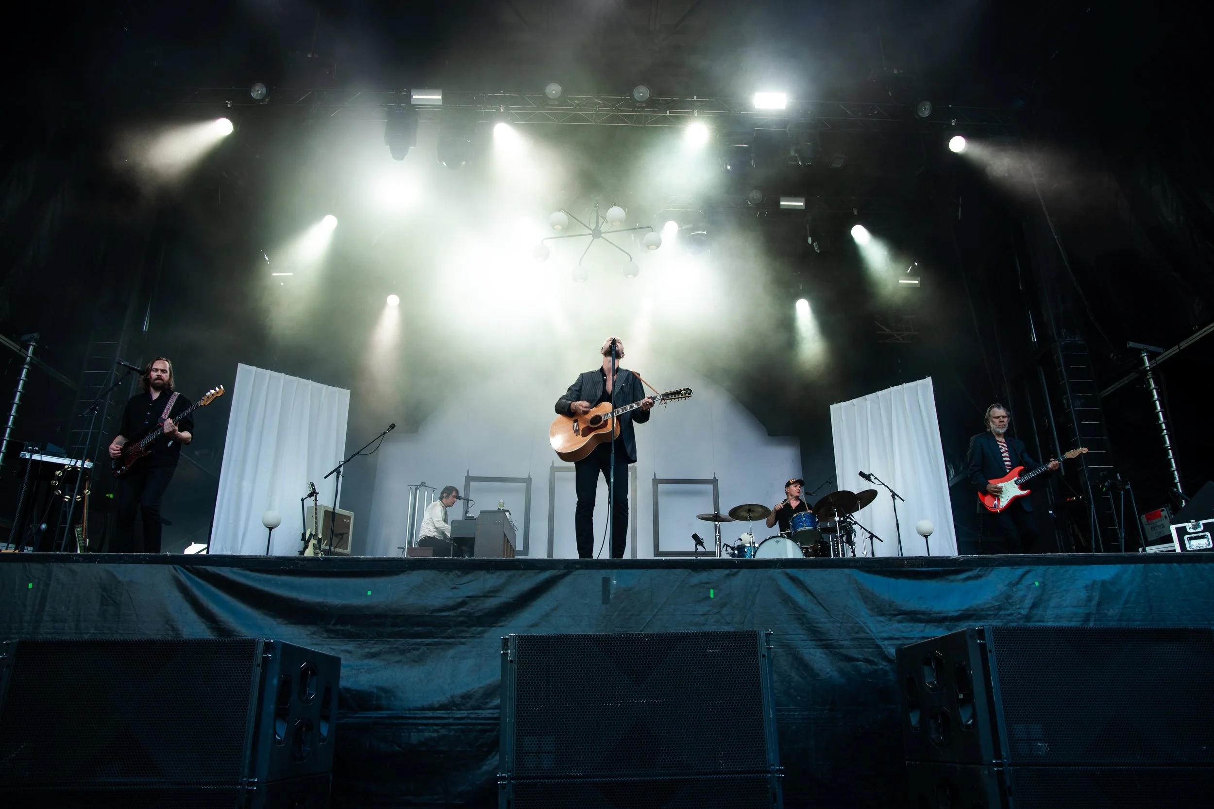 A band performs on stage with five members, including two guitarists, a drummer, a keyboardist, and a lead singer with an acoustic guitar. The stage is lit with bright lights and covered with white backdrop panels.