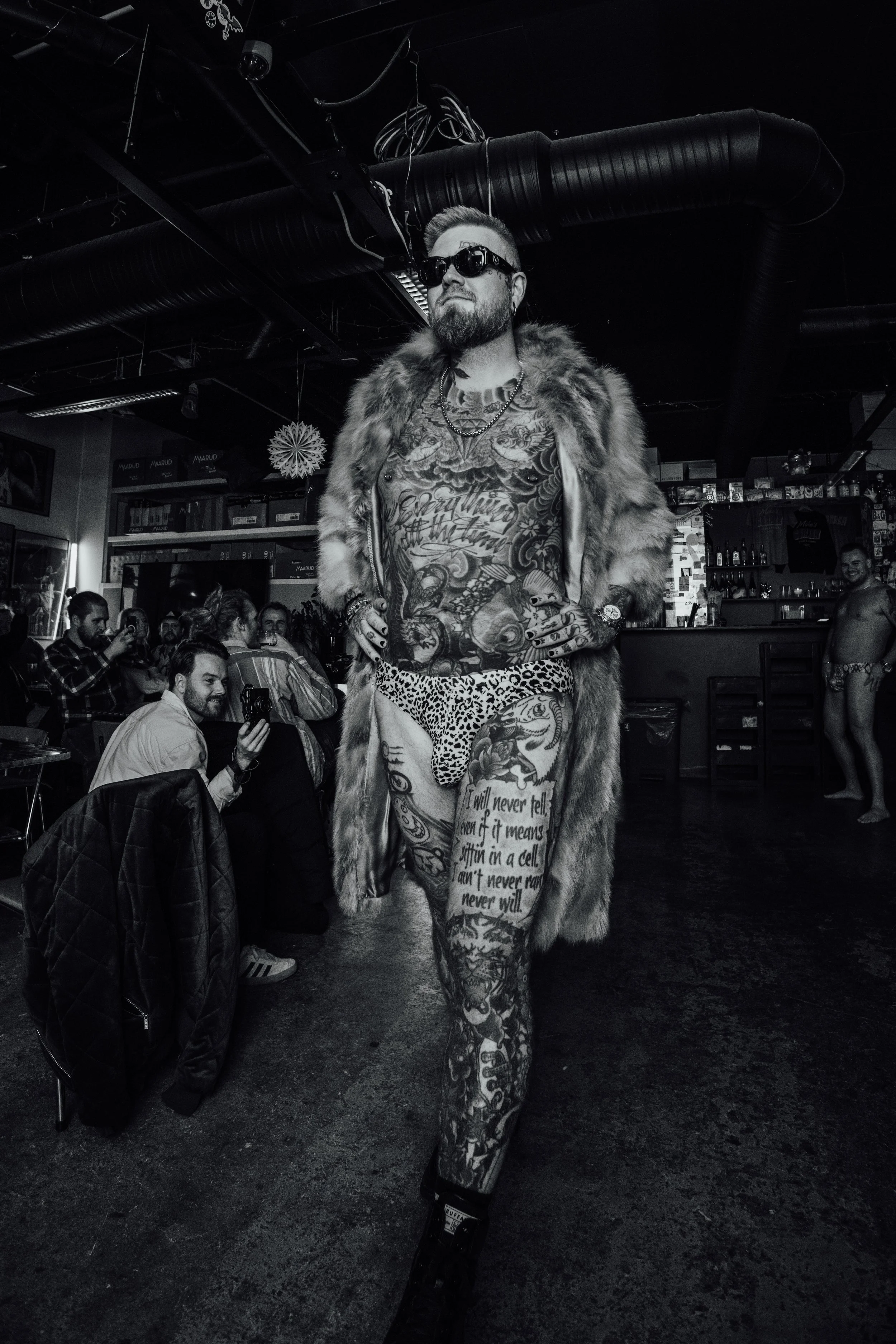 En fremtredende mann med mange tatoveringer, briller og pelsjakke går foran i en klubb, med andre mennesker som ser på og tar bilder. Skullet speedo lansering