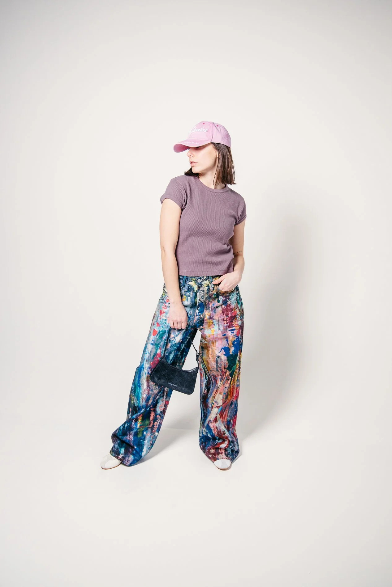 Kvinne i rosa baseball-cap, lilla t-skjorte og fargerike bukser, holder en liten svart veske, står mot en enkel lys bakgrunn.