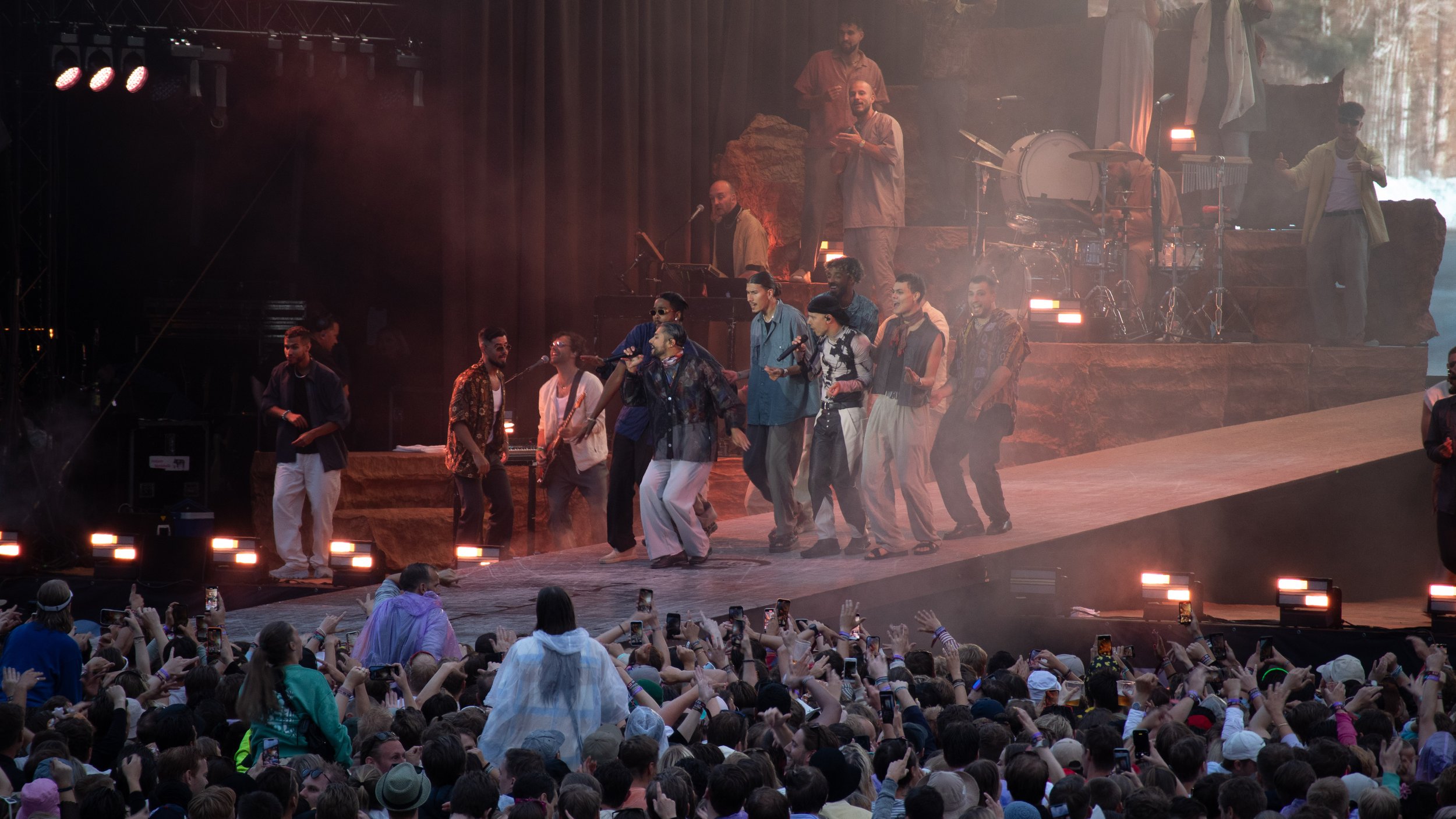 En stor gruppe musikere på scenen under en konsert, med en stor publikum foran, mange tar bilder med mobiltelefoner, og scenen er opplyst med varme lys. Karpe Slottsfjell 2022