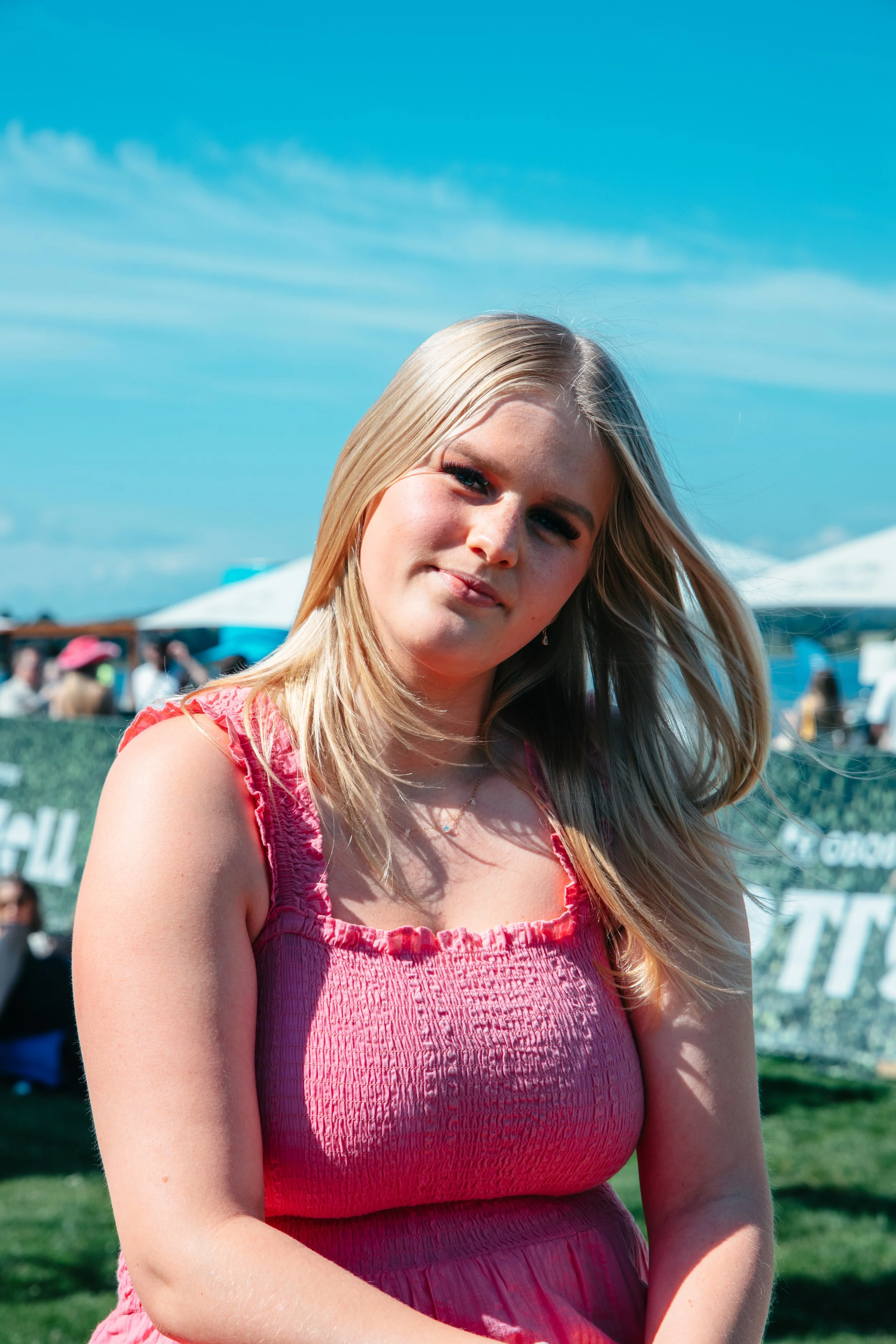 Porträtt av en ung kvinna med långt blond hår, som bär en rosa klänning, på en utomhusfestival eller marknad under en klar blå himmel. Slottsfjell Festivalen 2025