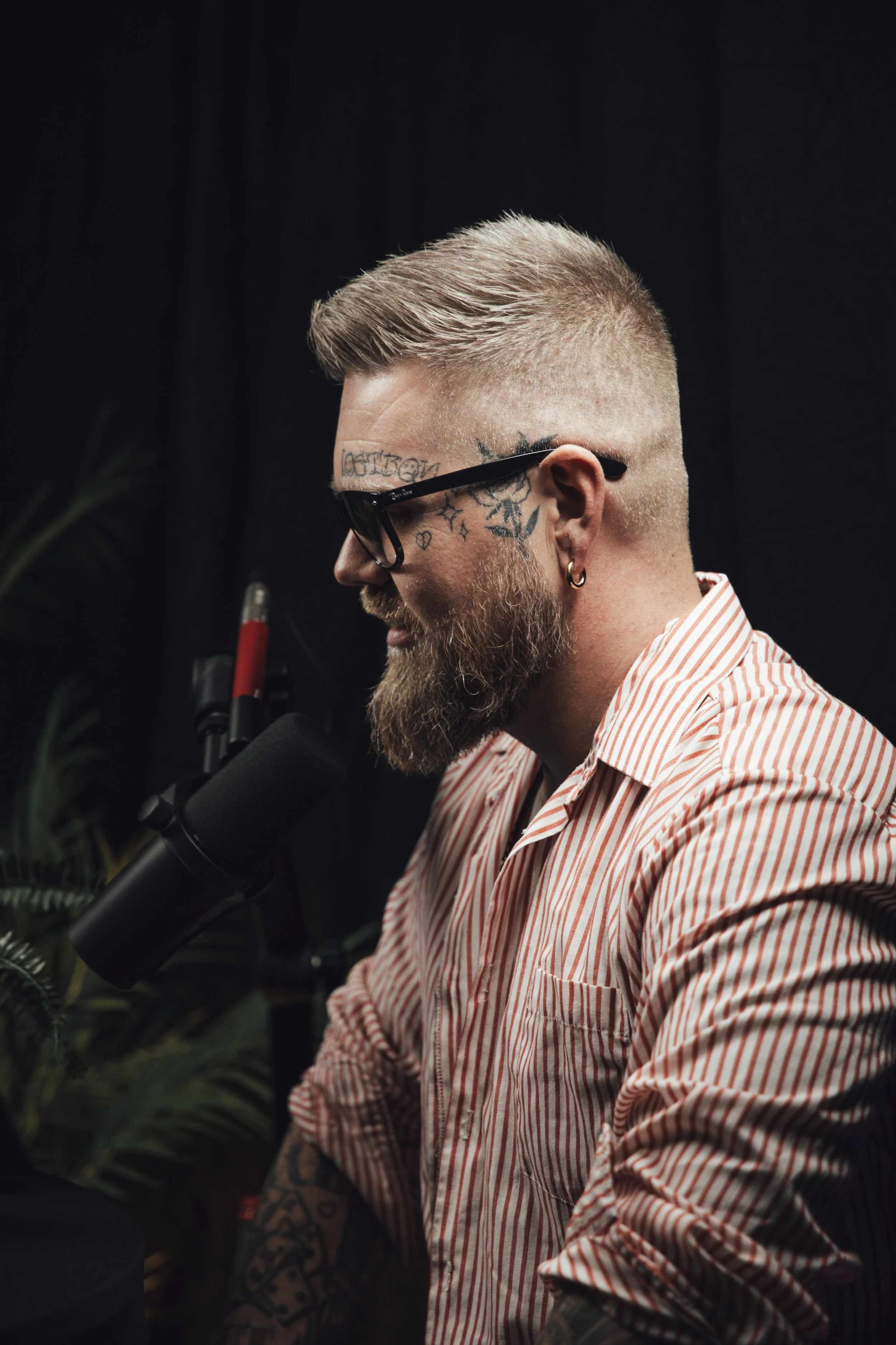 Profilbilde av en mann med tatoveringer, brunt skjegg, og kort, lyst hår, som snakker inn i en mikrofon.