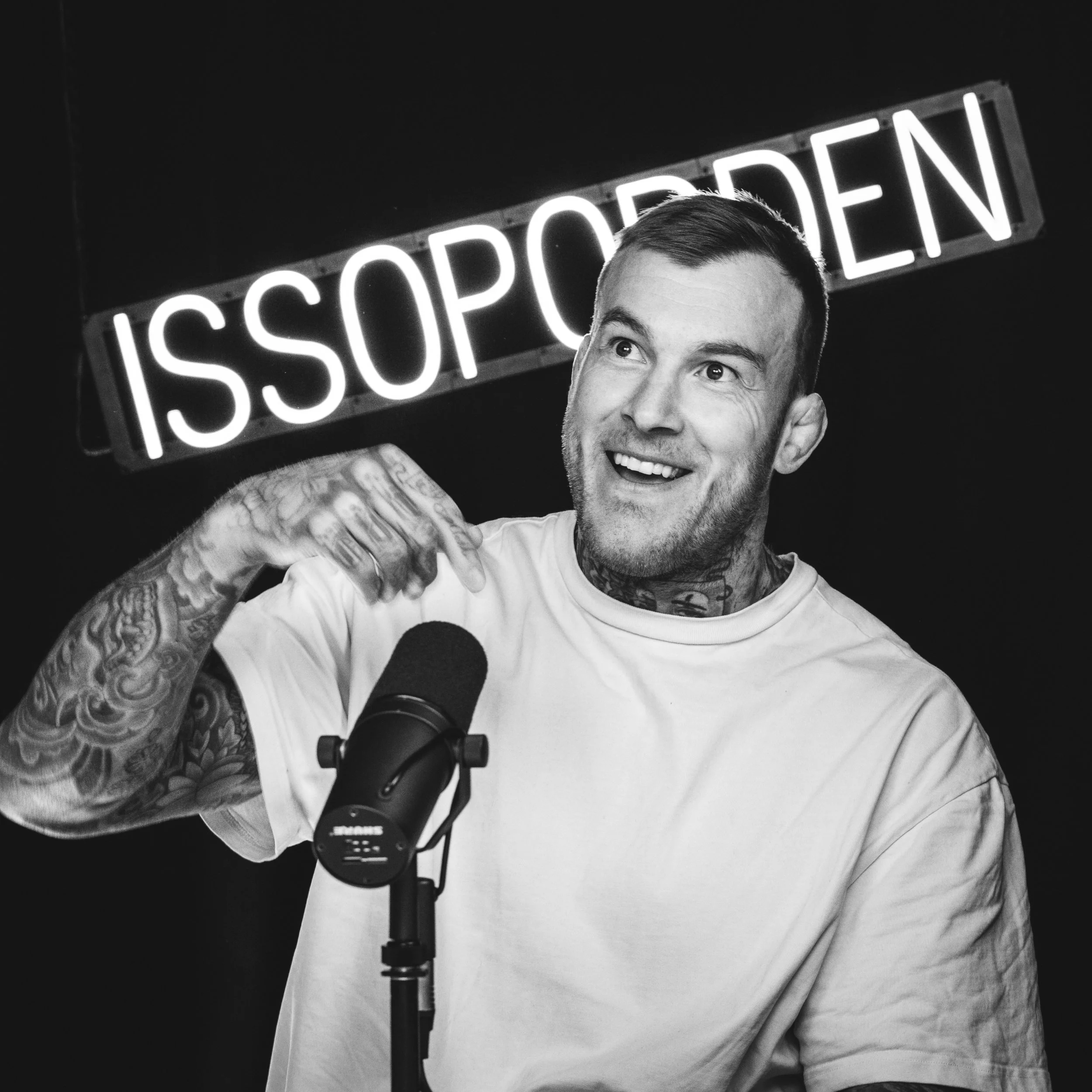 En mann med tatoveringer snakker inn i en mikrofon, med et opplyst neon-skilt i bakgrunnen som sier 'ISSOPPDEN'.