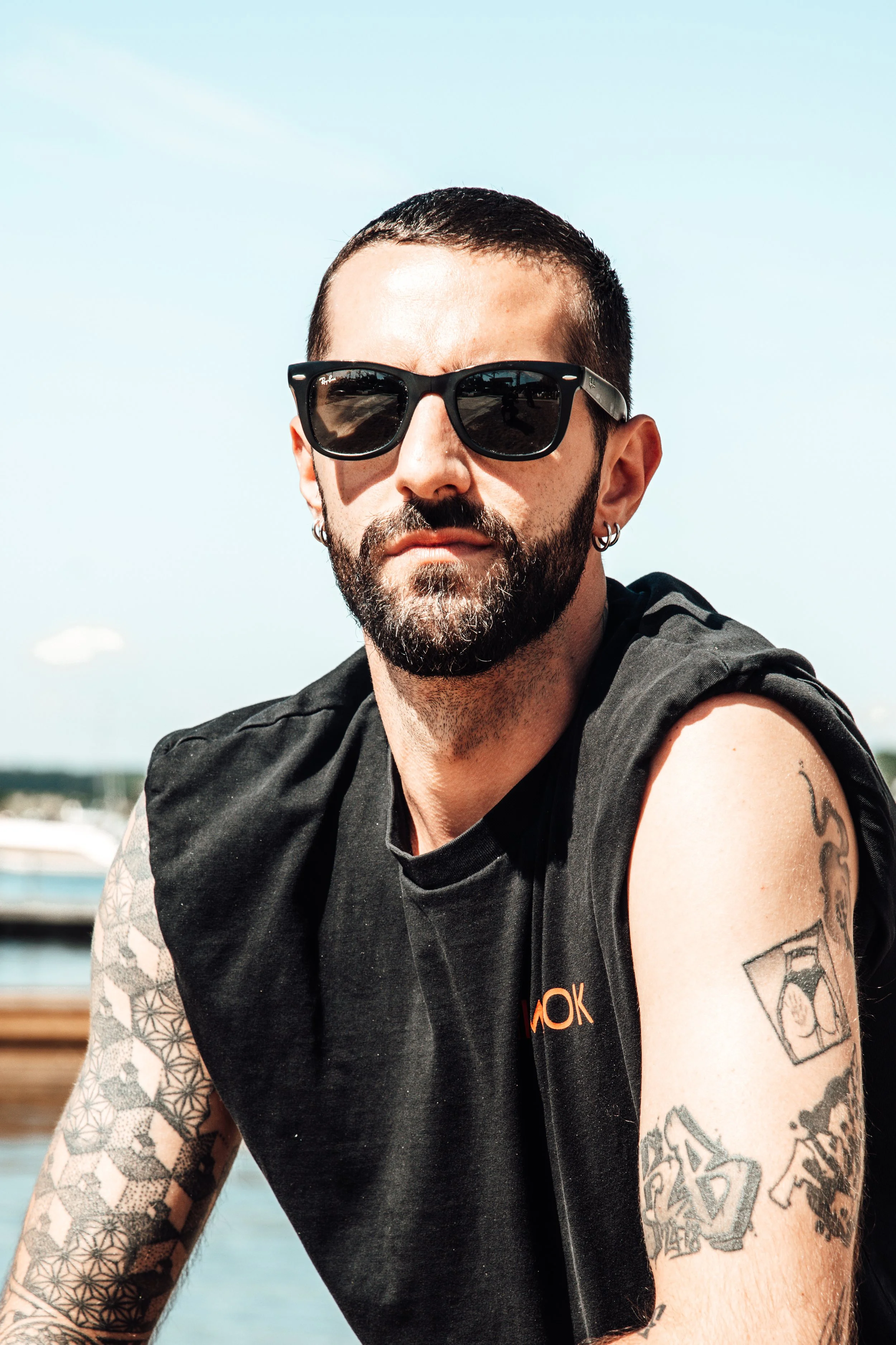 En mann med mørk skjegg, solbriller og tatoveringer på armene, utendørs med vann og himmel i bakgrunnen. Slottsfjell Festivalen 2025
