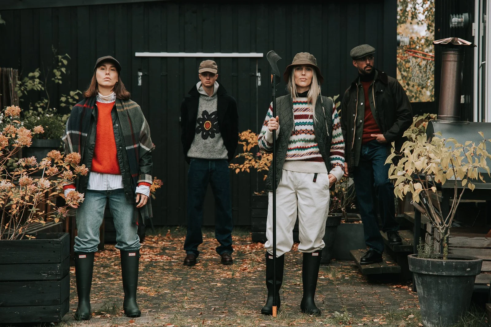 Five people standing outdoors in a garden, dressed in fall fashion with sweaters, jackets, and rubber boots, with plants and autumn leaves around them.