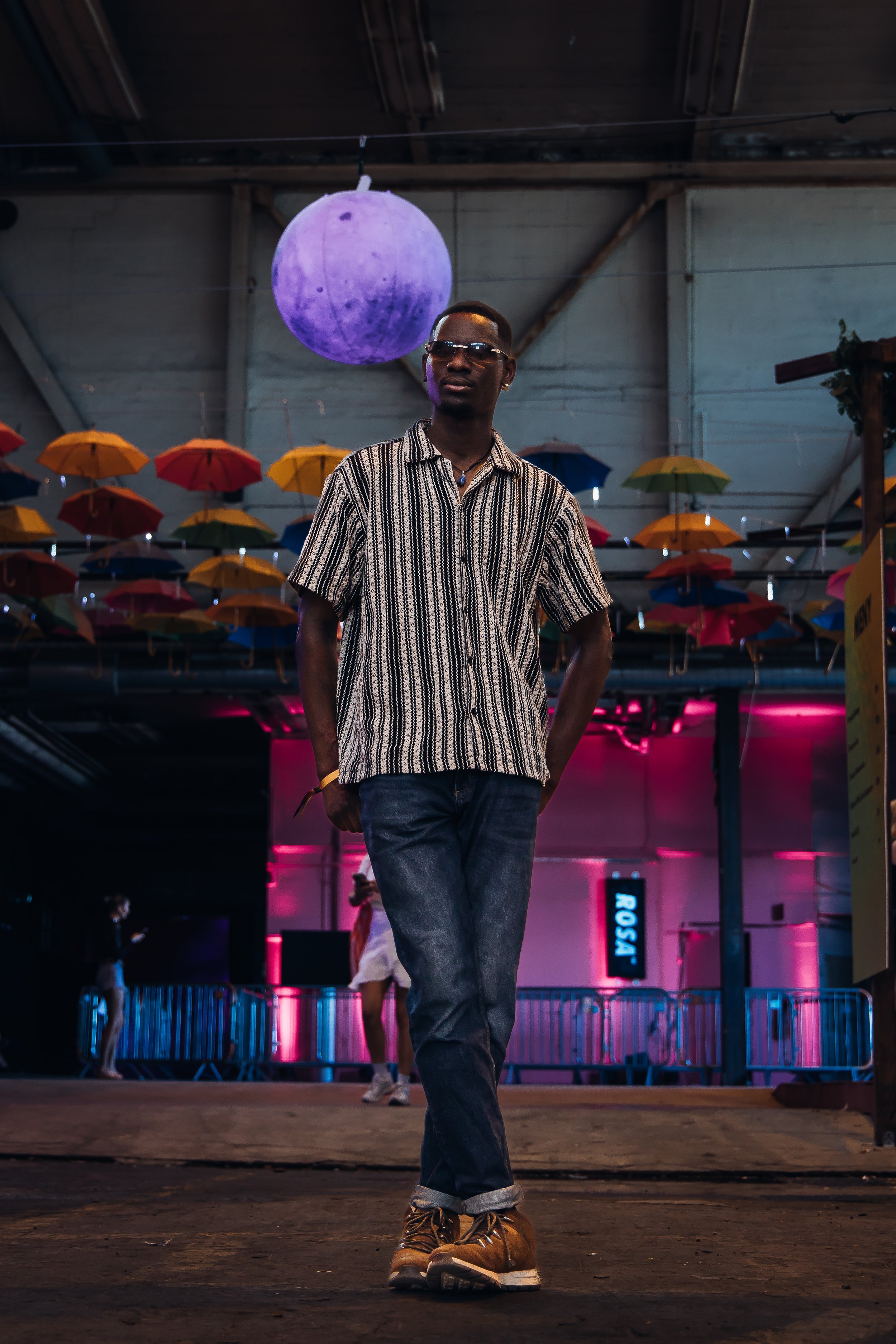 En ung mann med solbriller som går inn i et rom med fargerike paraplyer hengende fra taket og neonskilt i bakgrunnen.