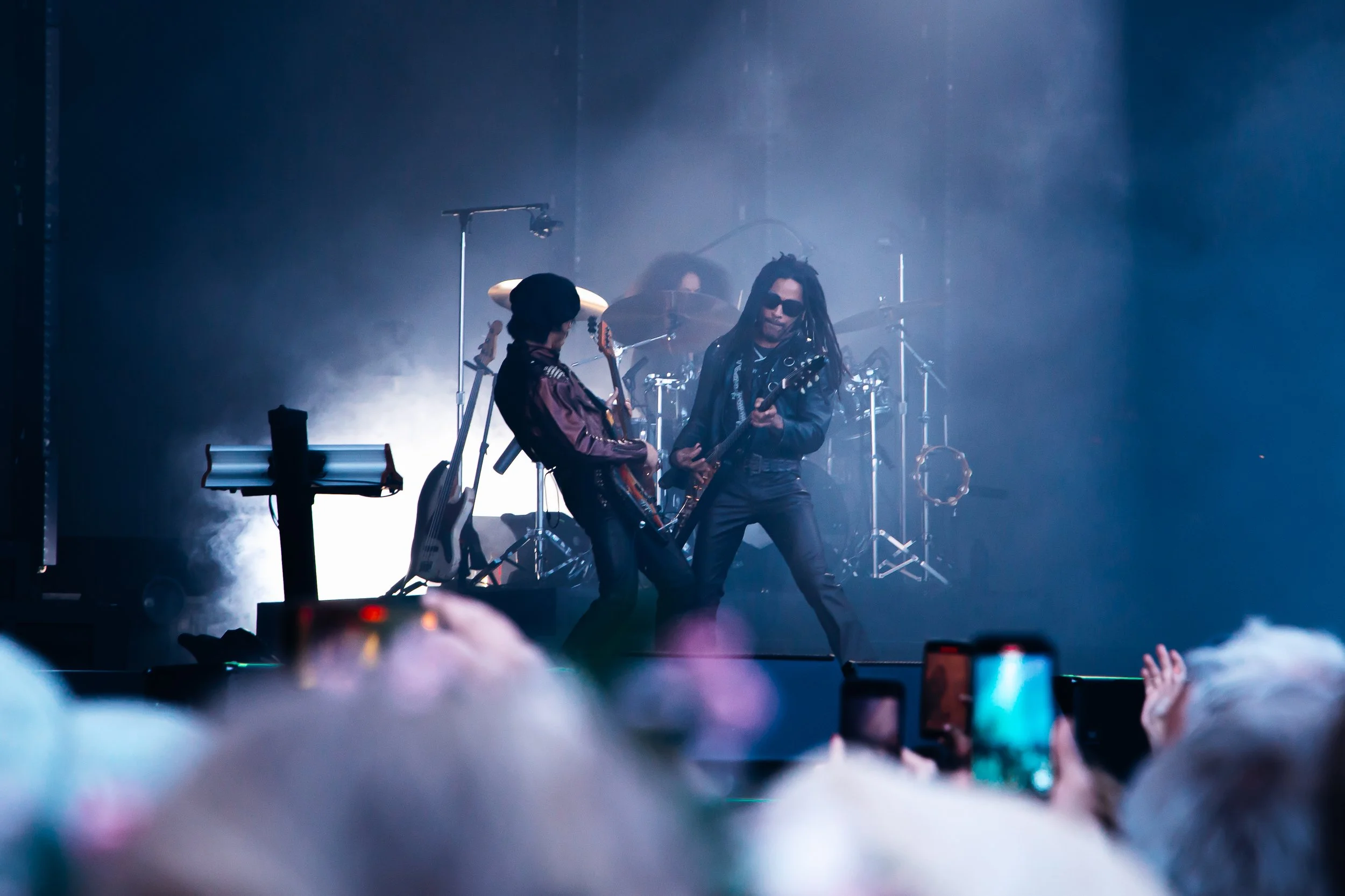 Two musicians performing on stage, playing electric guitars; one person with long dreadlocks and sunglasses, the other person with a wide-brim hat and a dark jacket. A drummer is visible in the background amid stage fog, with crowd members taking pho