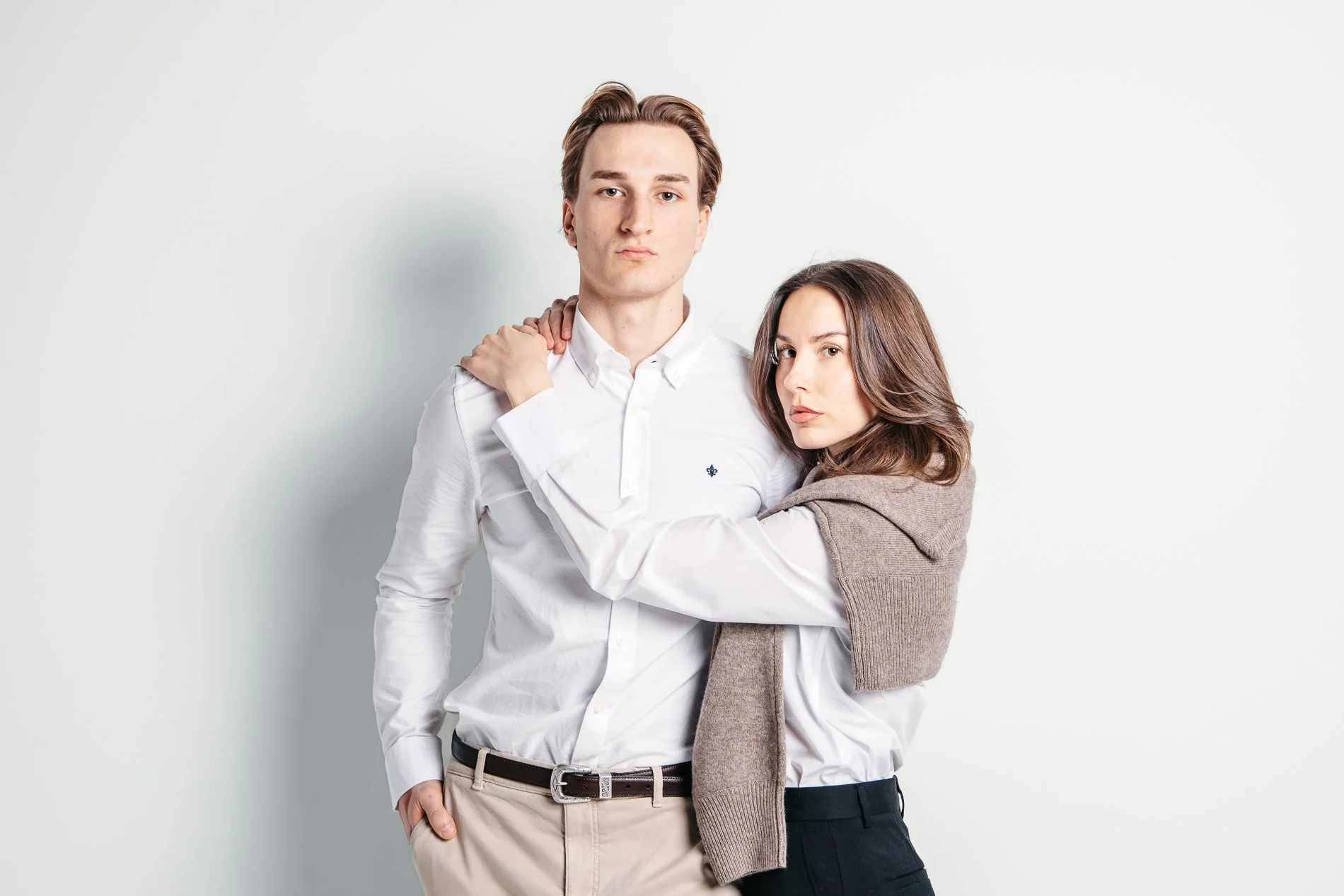 En ung mann og en ung kvinne poserer mot en lys bakgrunn, kledd i hvit skjorte og svart bukse, med kvinnens arm rundt mannens skuldre.