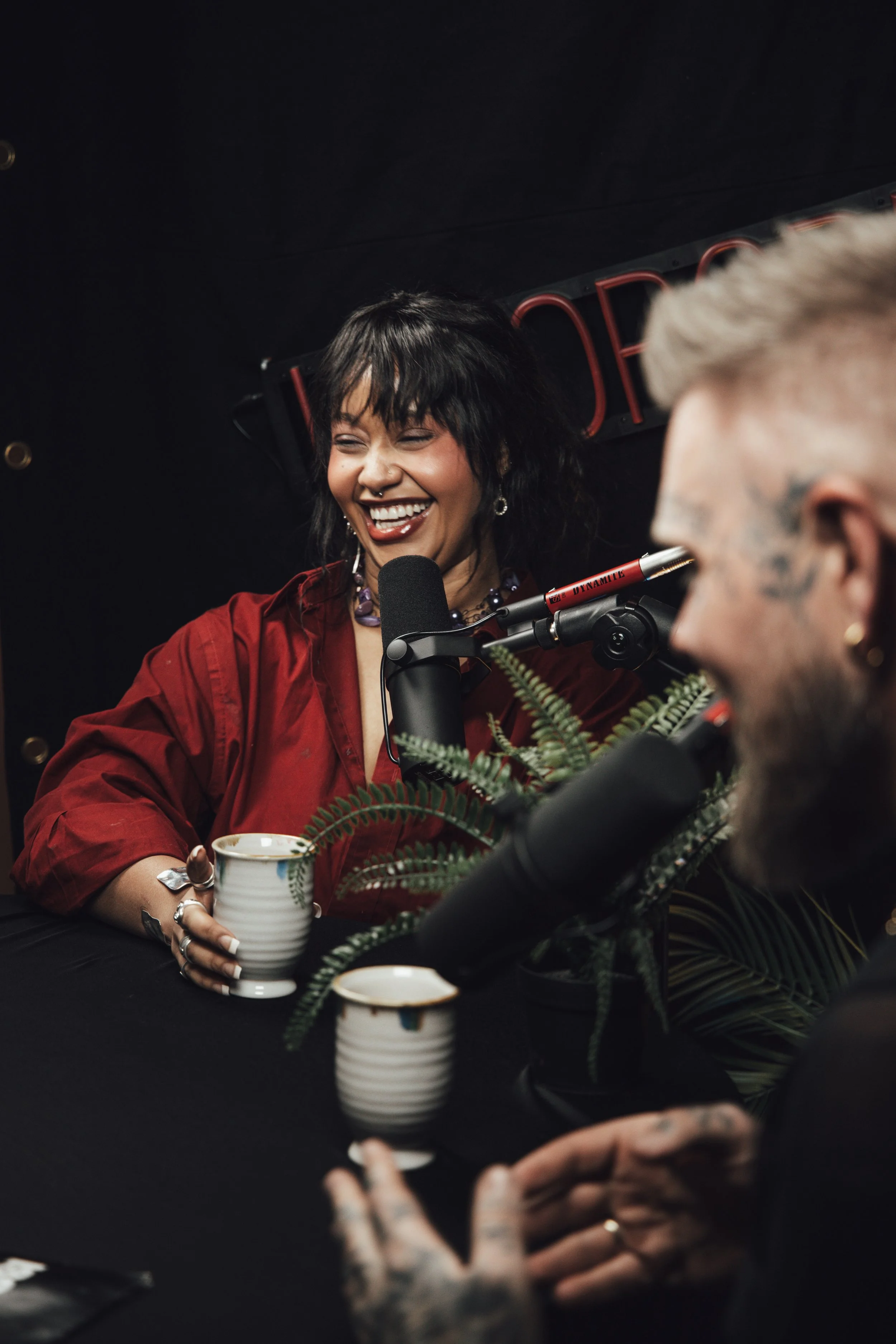 To personer snakker i en mikrofon under en radiopodcast, kvinnen smiler og har kort svart hår og er kledd i en rød skjorte, mannen har utstående tatoveringer og blond hår.