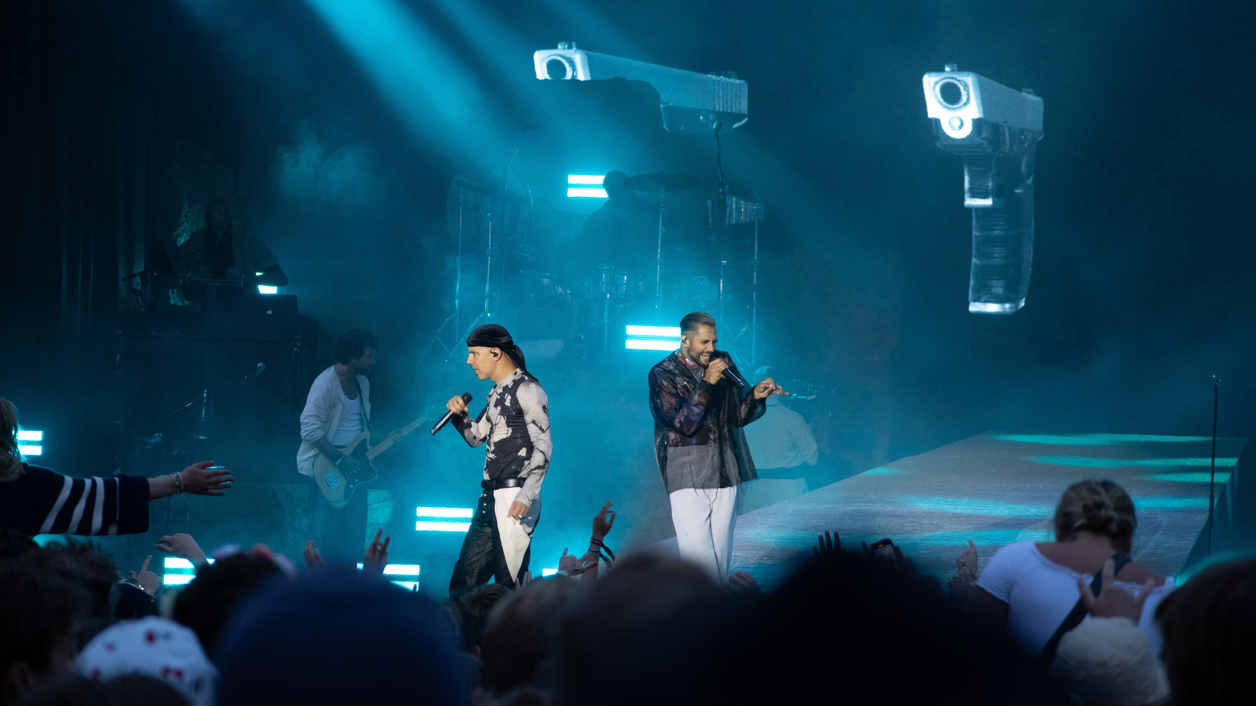 En artistgruppe oppfører seg på en scene under en konsert, med to sangere foran og et band i bakgrunnen, omgitt av blå lys. Karpe Slottsfjell 2022