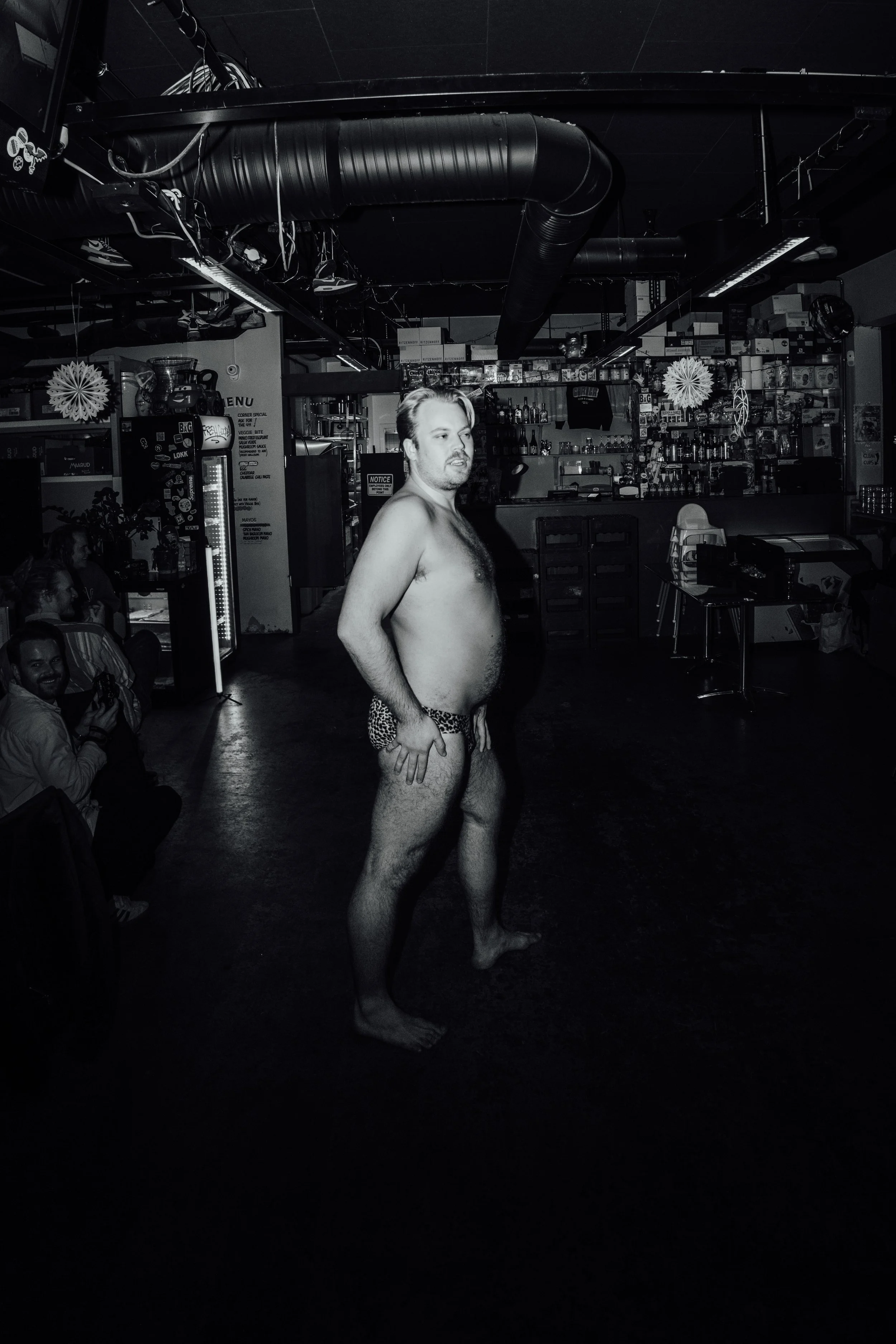 En mann i baren, iført bare undertøy, står i midten av rommet, omgitt av andre personer som sitter og ser på.