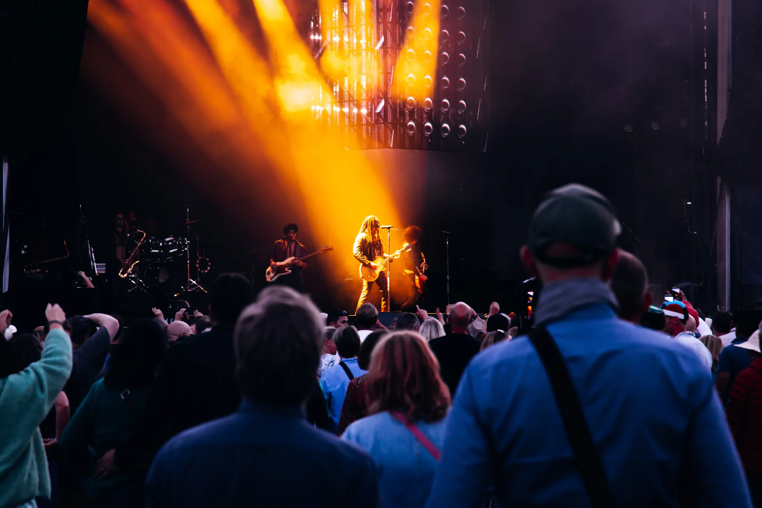 A live concert with a band performing on stage illuminated by yellow and orange lights. The audience is watching, some taking photos.