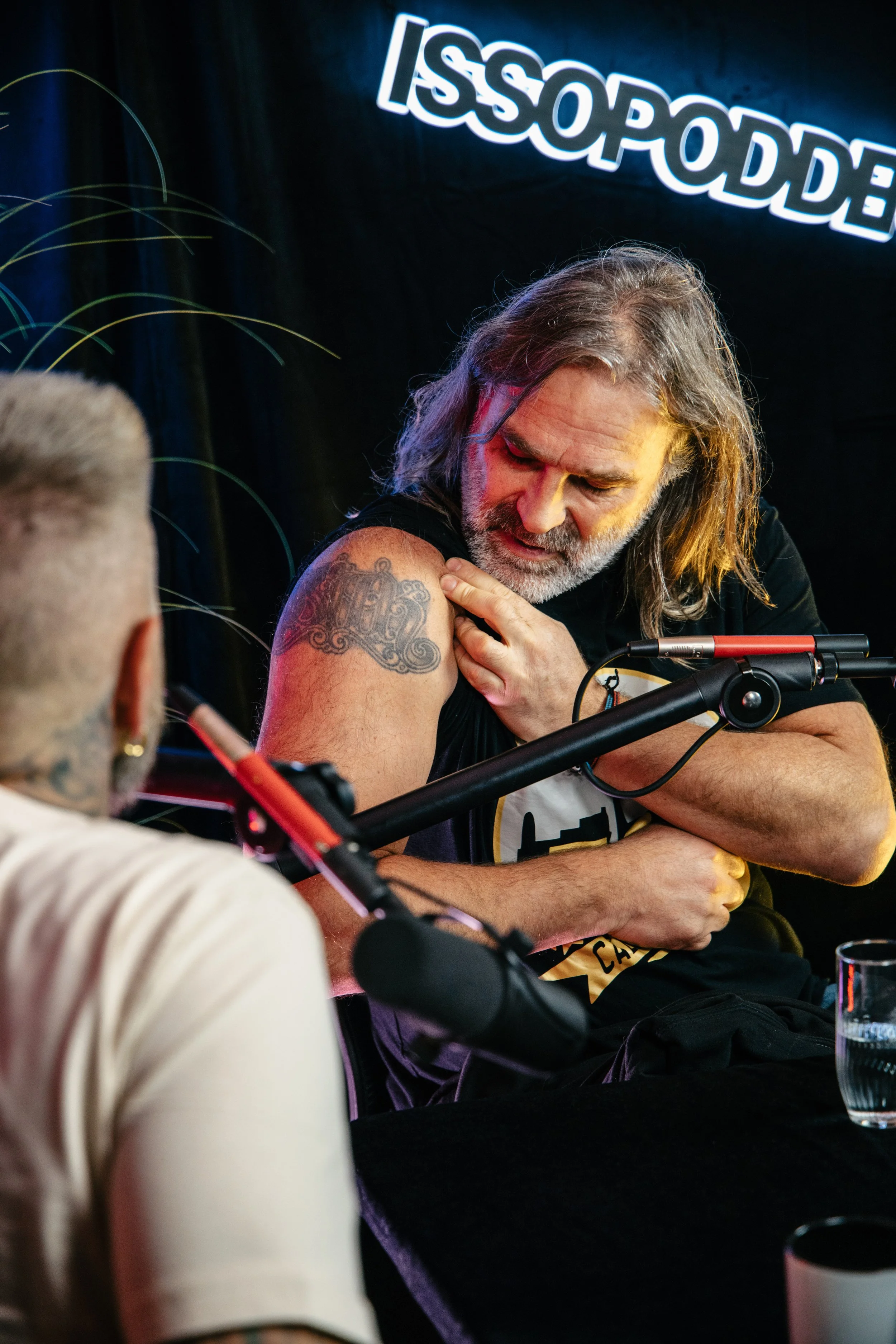 En eldre mann med langt grått hår og skjegg viser opp armen sin med en tatovering under en podcastpilot. Han ser på tattooen med interesse, mens en annen person med kort hår og tattooer er delvis synlig i forgrunnen.