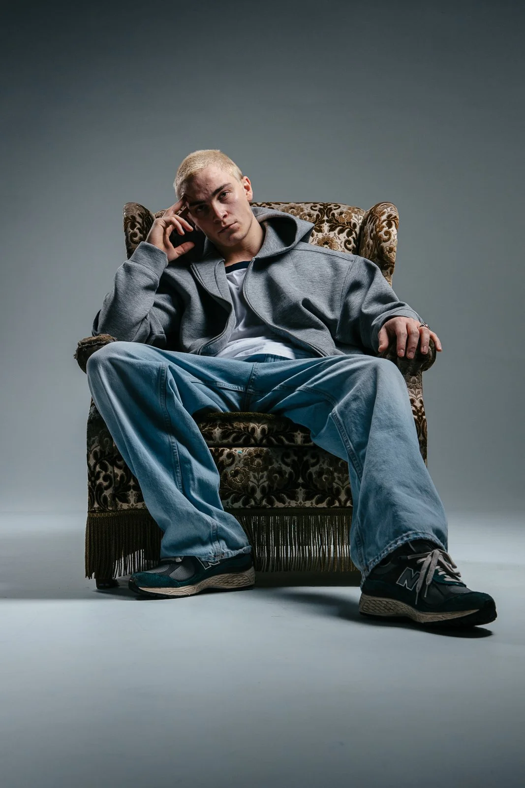 Teenaged young male with blond hair sitting in a vintage armchair, wearing a gray hoodie and jeans, with a serious expression.
