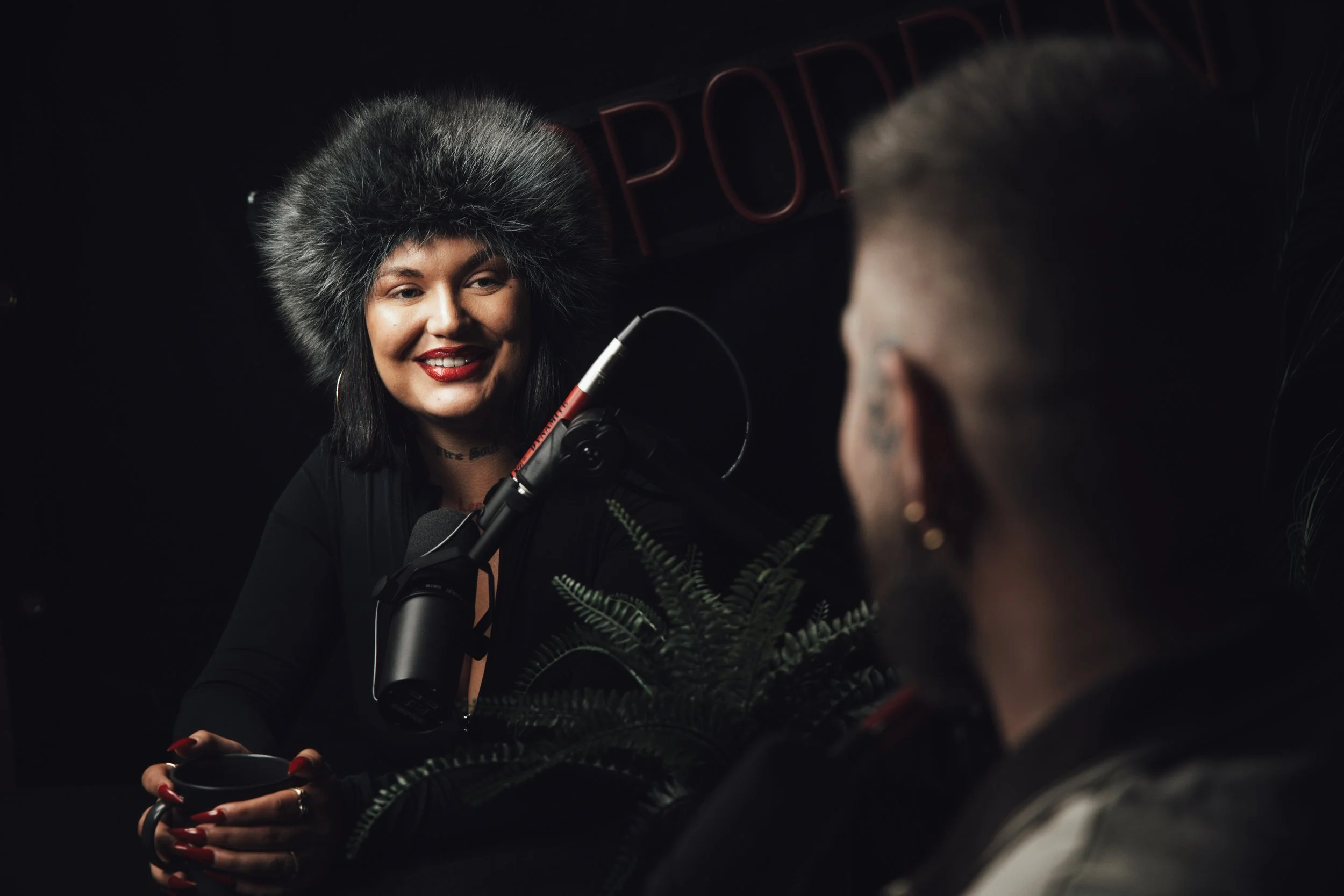 En kvinne smiler mens hun snakker i en mikrofon, det er en annen person i forgrunnen med ryggen mot kamera, utenfor fokus. Bakgrunn er svart med en delvis synlig neonskrift.