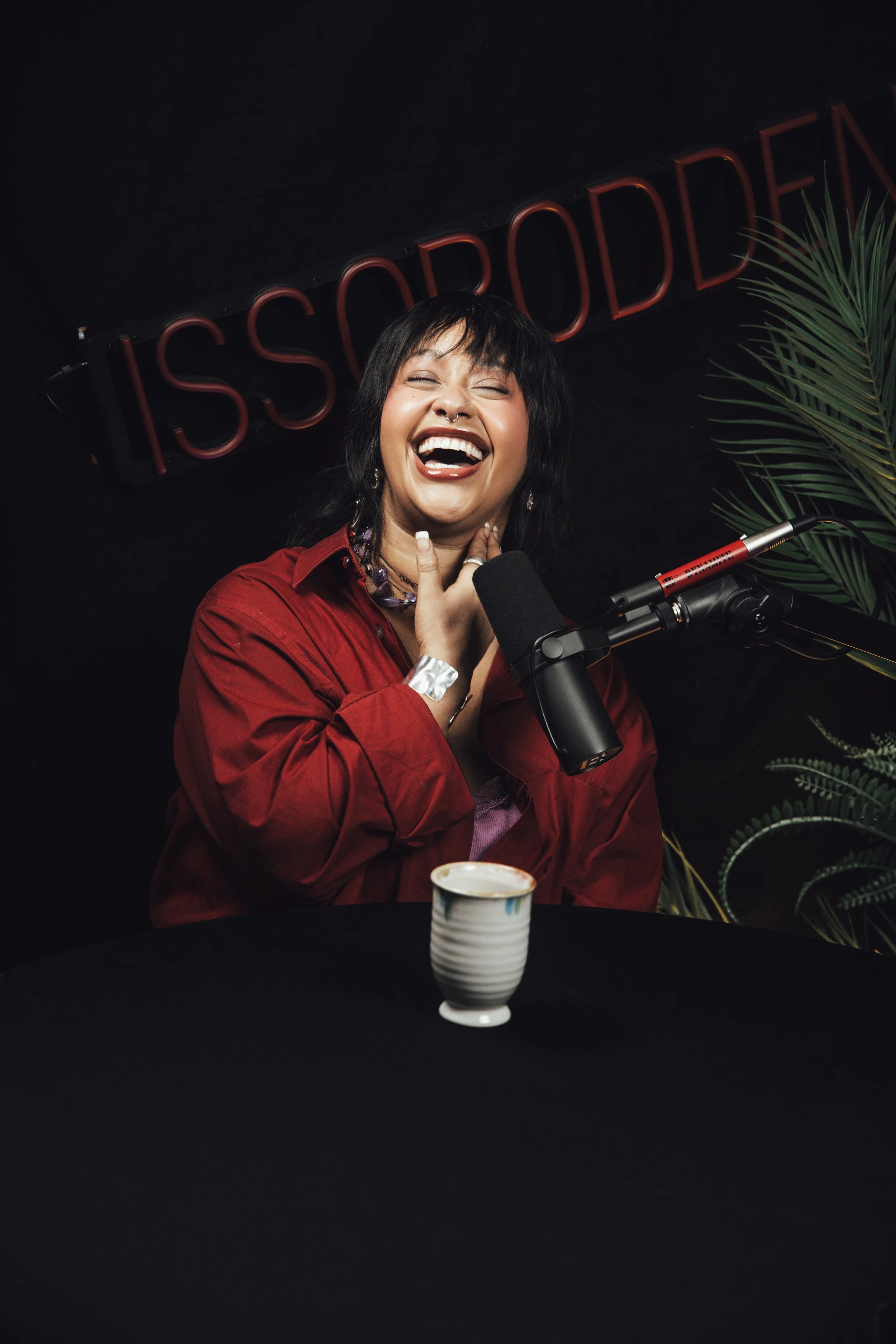 En kvinne ler mens hun snakker i en mikrofon ved et bord, med en kopp foran henne og en plante i bakgrunnen. Neonlys som sier 'ISSOU PODDEN' er synlig i bakgrunnen.