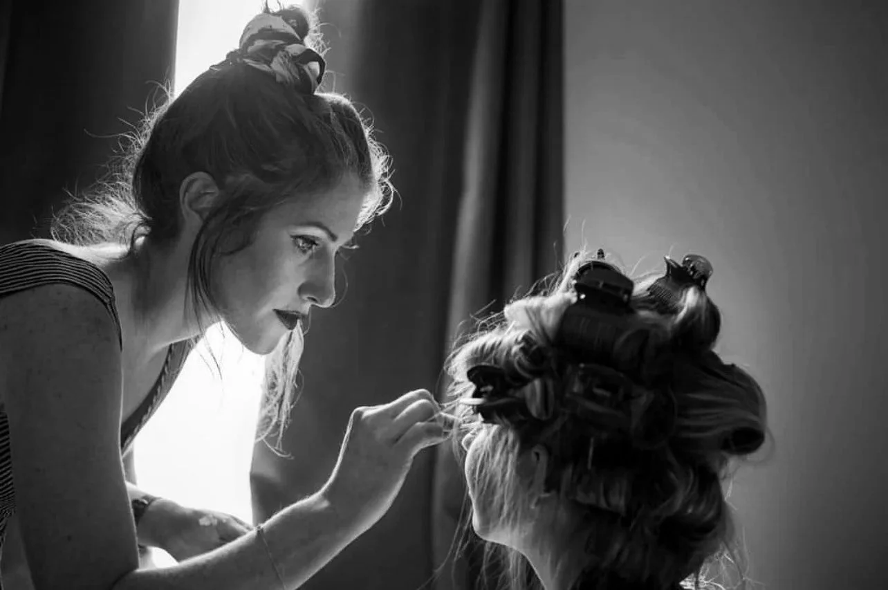 A woman with curlers in her hair is getting her makeup done by another woman with hair in a bun, both in a room with curtains and bright backlighting.