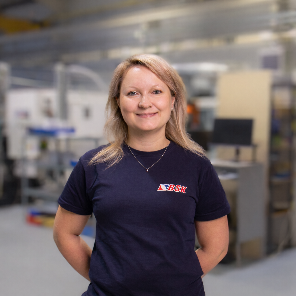 Frau in dunklem T-Shirt mit BSK-Logo in einer Werkstatt oder Fabrikhalle, lächelnd.