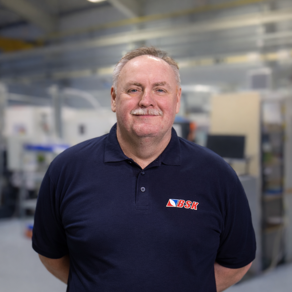 Ein Mann mit grauem Haar und Schnurrbart trägt ein dunkles T-Shirt mit dem Logo BSK, steht in einem Industrie- oder Bürogebäude.