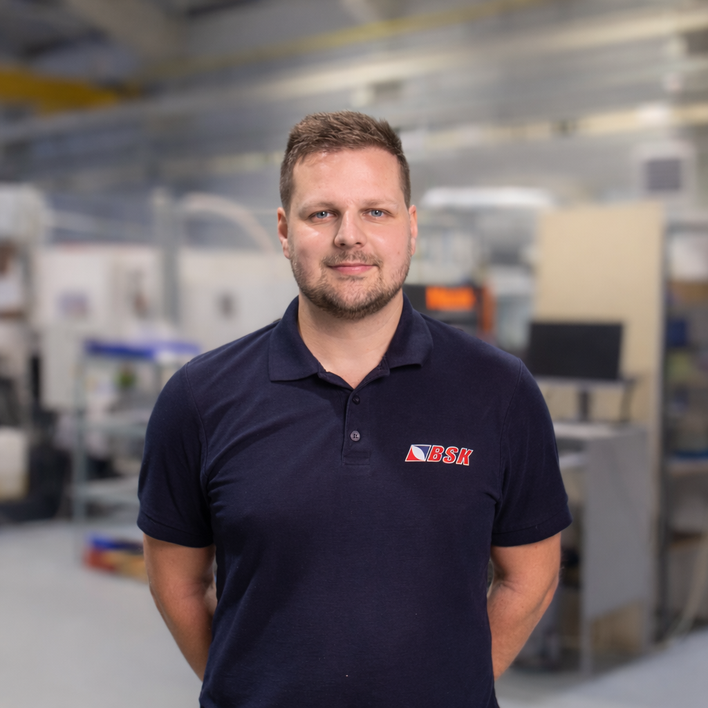 Mann in dunklem Polo-Shirt mit BSK-Logo steht vor industrieller Hintergrund in Werkstatt oder Lagerhaus.