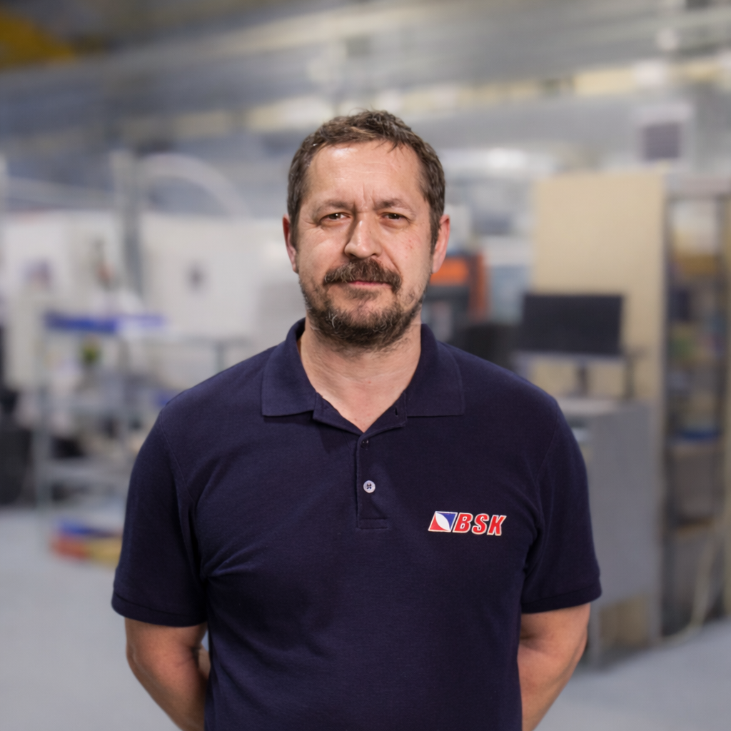 Ein Mann mit braunem Haar und Bart in einem blauen Polo-Shirt mit LOGO steht in einer industriellen Umgebung.