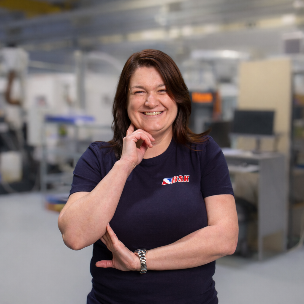 Frau mit dunklem T-Shirt und BSK-Logo, lachend, in einer technischen oder industriellen Umgebung.