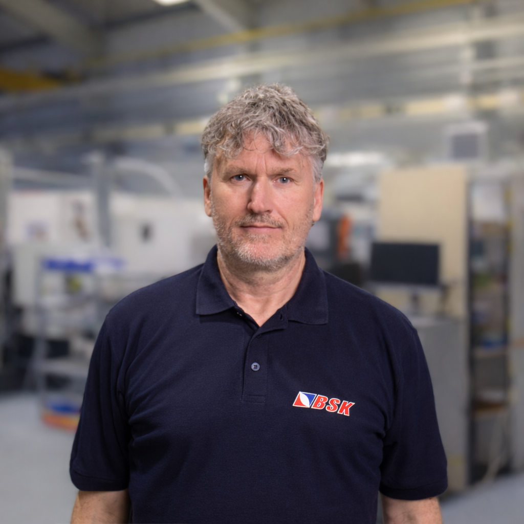 Ein Mann trägt ein schwarzes Polo-Shirt mit dem Logo 'BSK' in einem industriellen Umfeld.