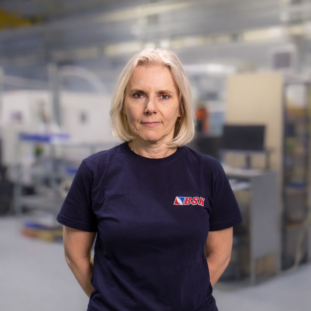 Eine blonde Frau trägt ein schwarzes T-Shirt mit dem Logo 'BSK' in einem Industrie- oder Laborumfeld.
