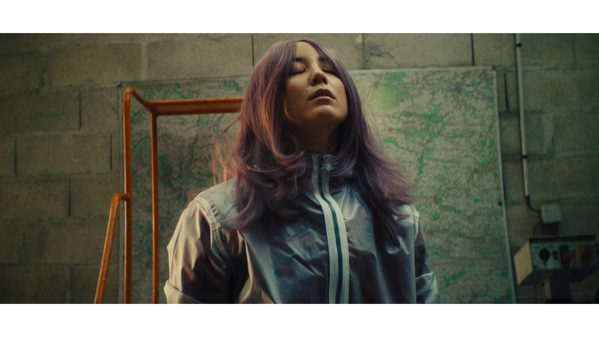 A woman with purple hair and closed eyes stands in front of a wall with a map, wearing a shiny gray jacket, with a worried expression.
