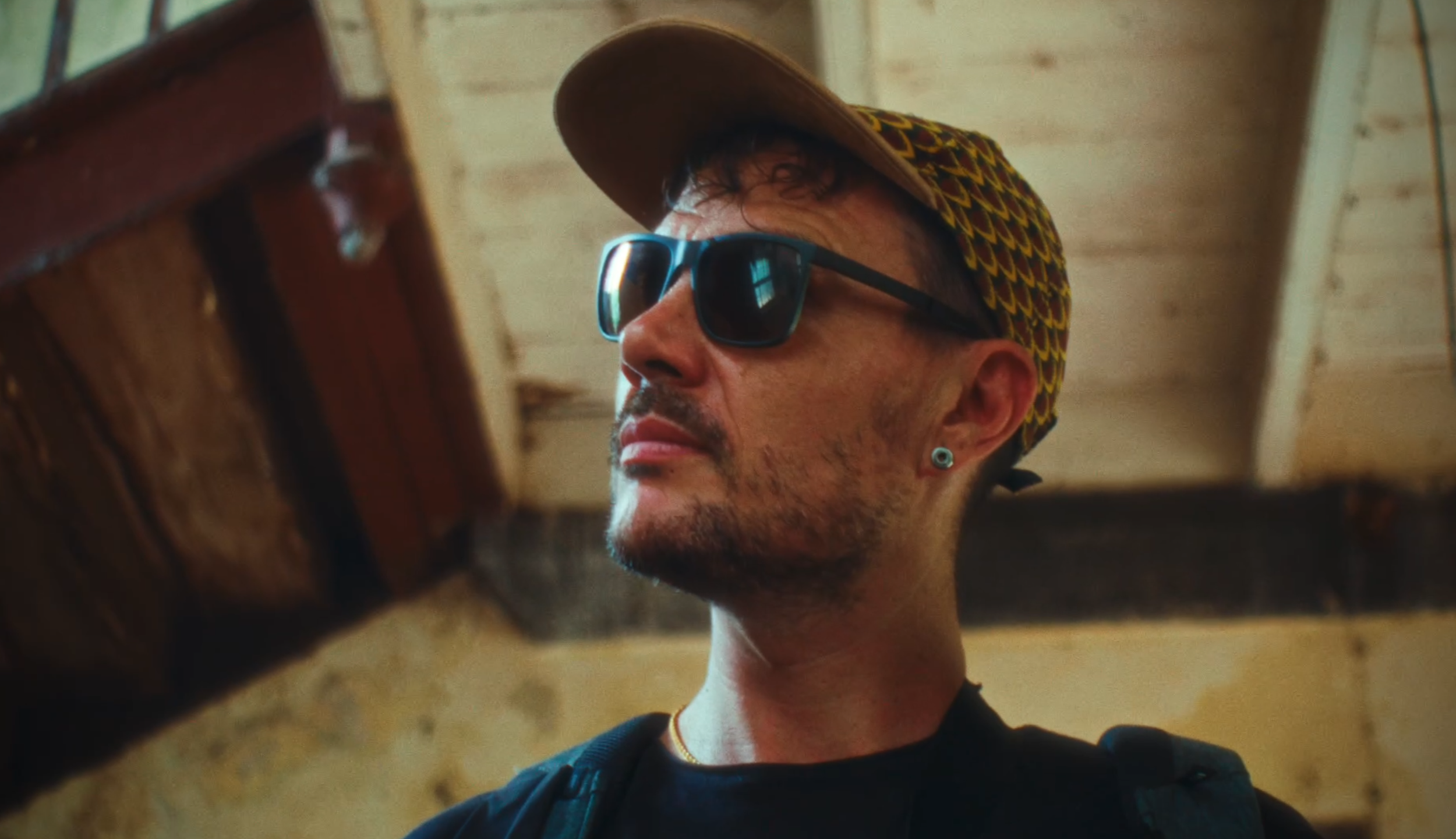 A man wearing a baseball cap, sunglasses, and an earring, looking at a wooden object in a rustic setting.