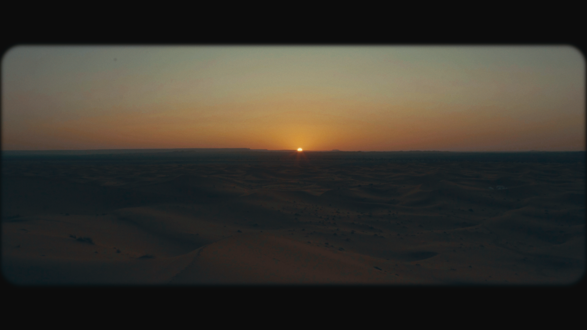 Sunset over a desert landscape, with sand dunes extending into the horizon under a clear sky.