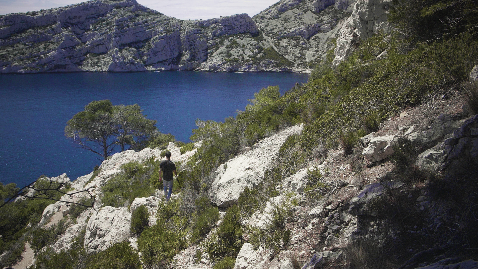 A person walking along a rocky hillside with a view of a large blue lake surrounded by rocky cliffs and green vegetation.
