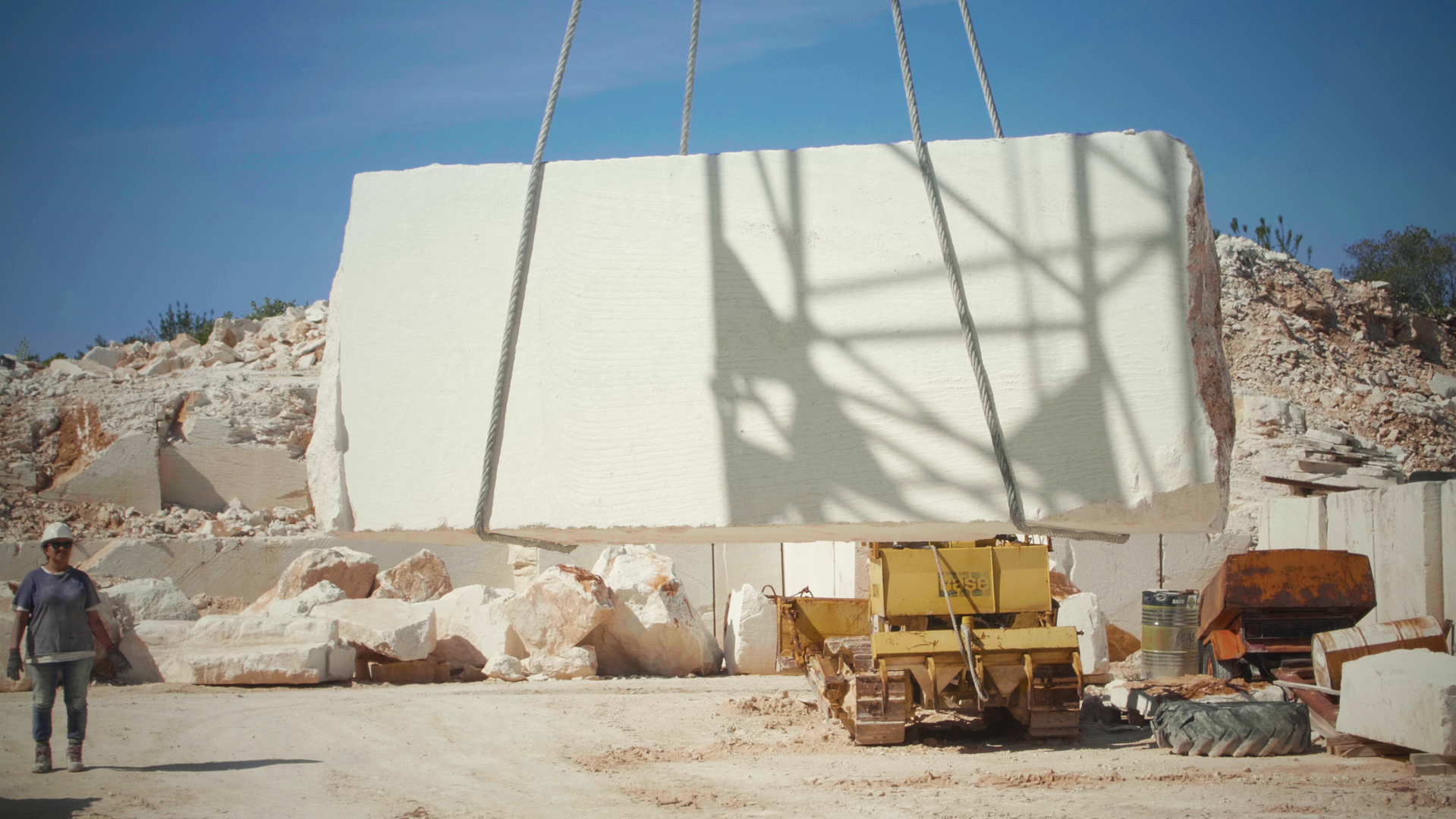 A large white marble slab is lifted by a yellow crane at a quarry, with workers nearby and a clear blue sky above.