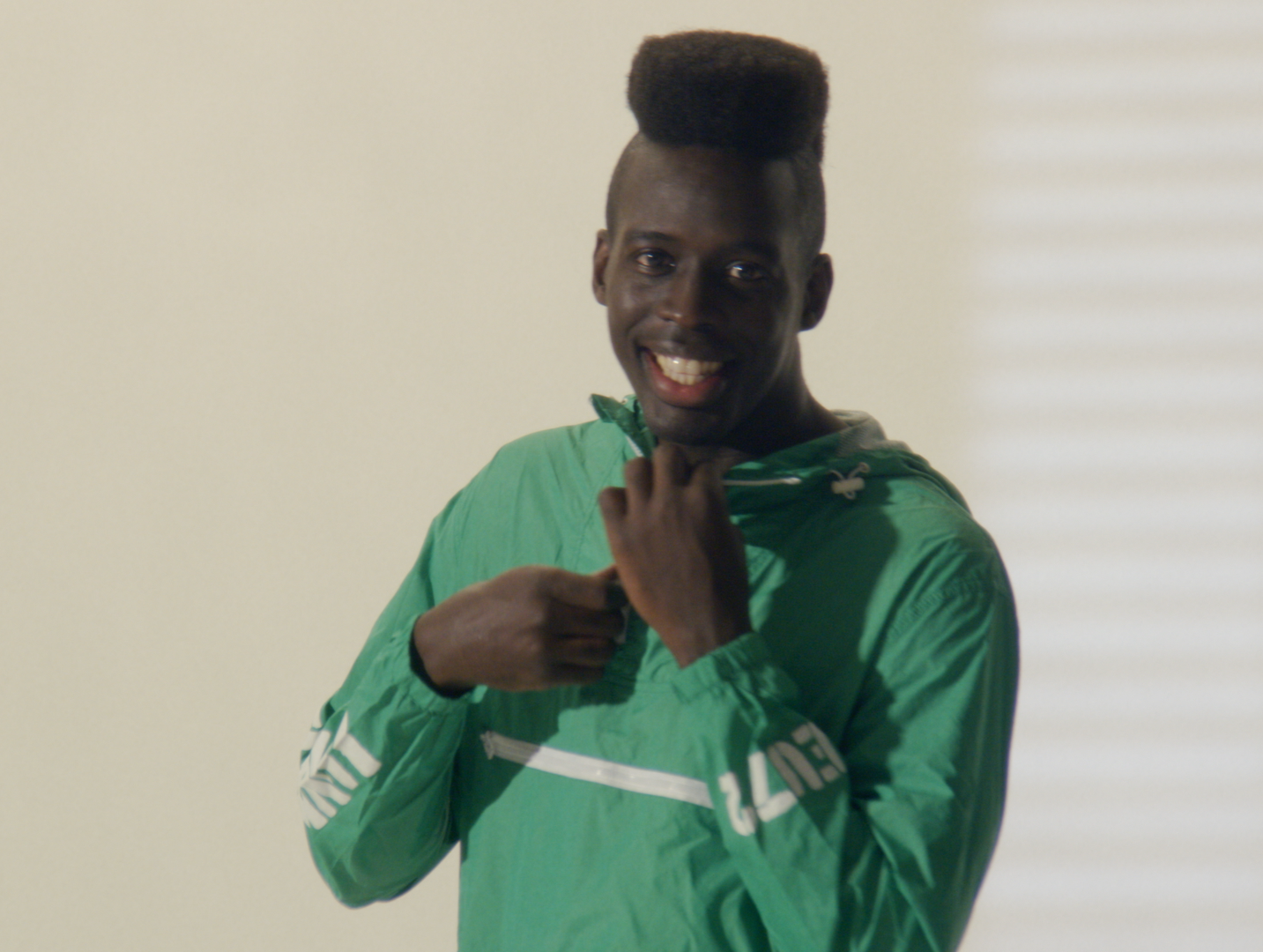 A young man with an upward hairstyle smiling and adjusting his green jacket.