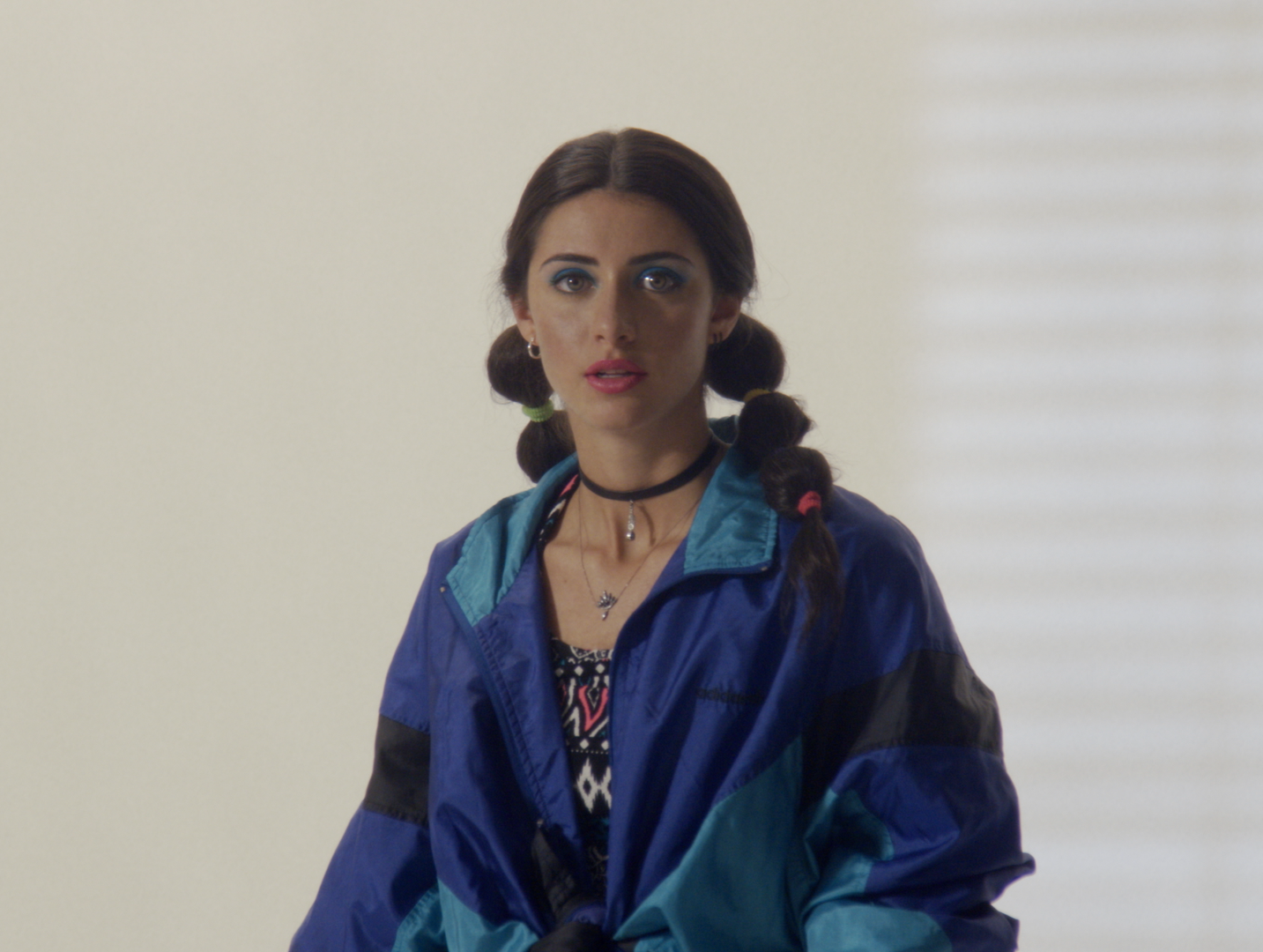 Woman with dark hair in pigtails wearing colorful 90s-style jacket and jewelry, standing indoors with window blinds in background.