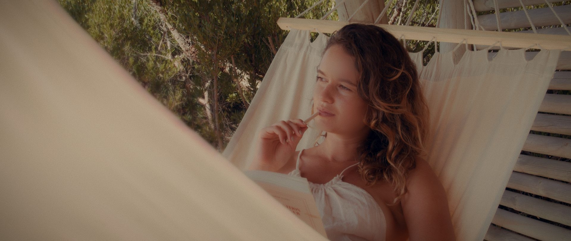 Young woman with curly hair reclining on a hammock outdoors, holding a book in one hand and a popsicle in the other, with trees and wooden fence in the background.