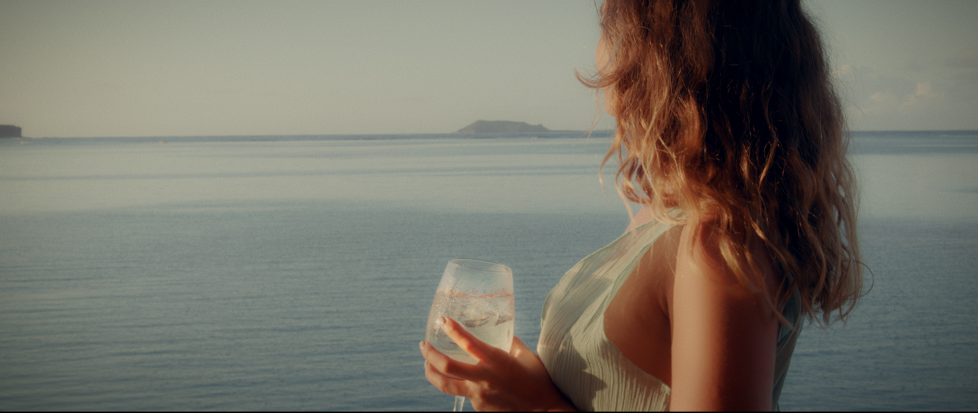 A woman with curly hair holding a glass of water by the ocean with a distant island in the background.