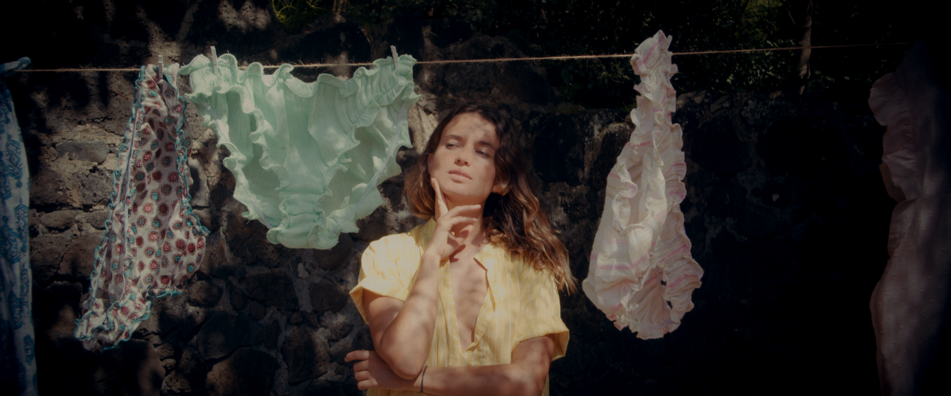 A woman with long wavy hair in a yellow shirt standing in front of a stone wall with colorful laundry hanging on a line.''
