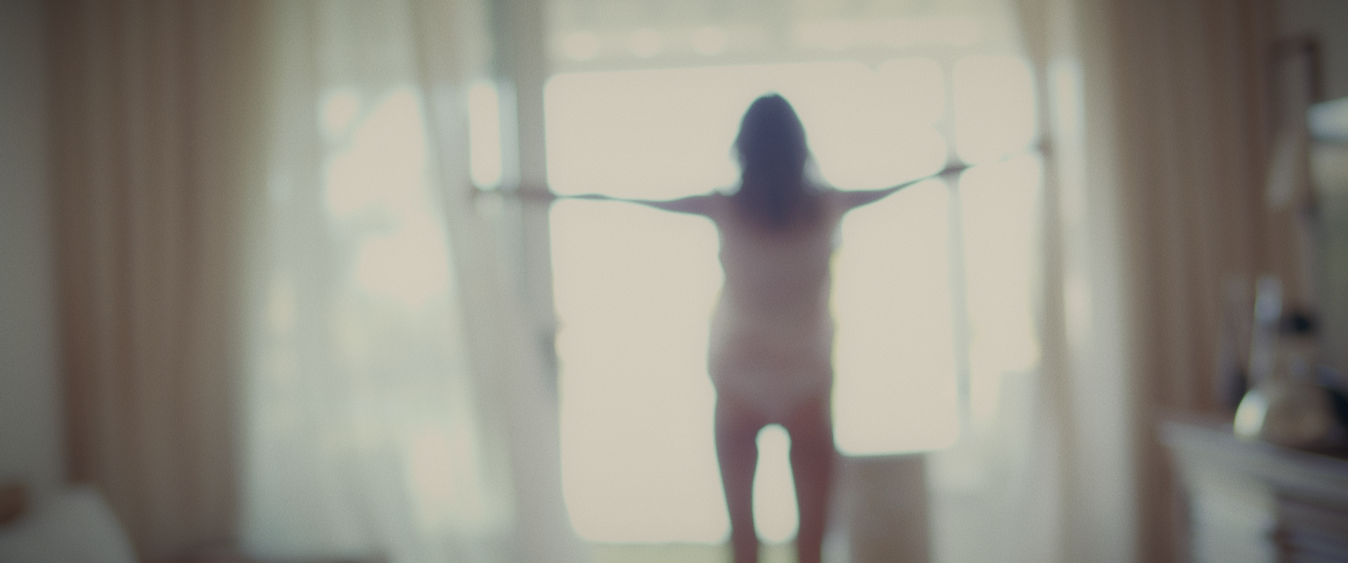 A woman standing by a large window with her arms outstretched, silhouetted against bright sunlight in a room.