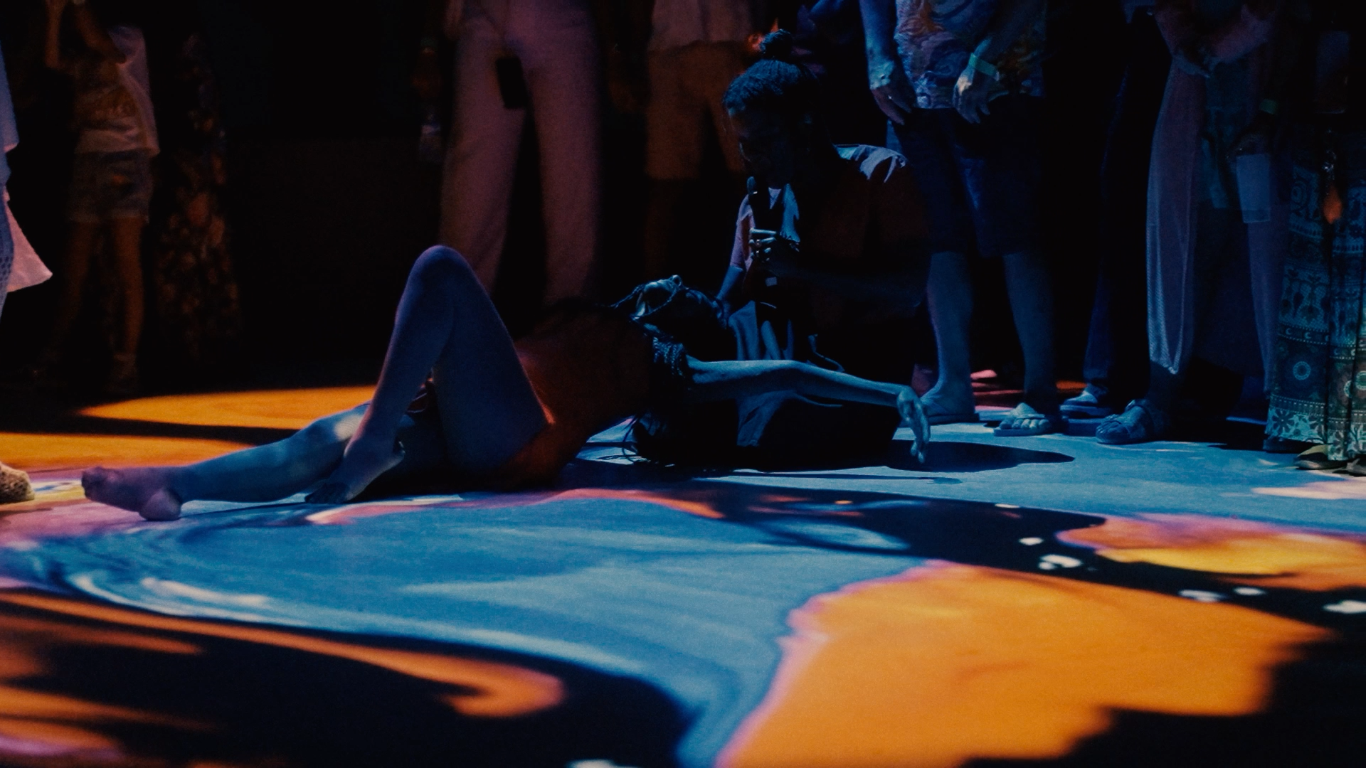 A woman is lying on the floor with her back arched, surrounded by a group of people standing and watching in the dark. The scene is illuminated by colorful lighting.