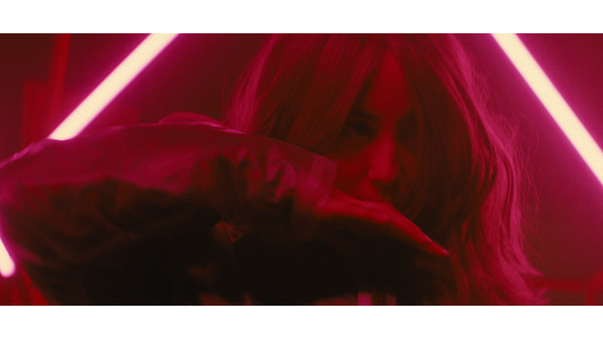 A woman with long hair partially covering her face, illuminated by pink neon lights, holding an umbrella in a dark setting.