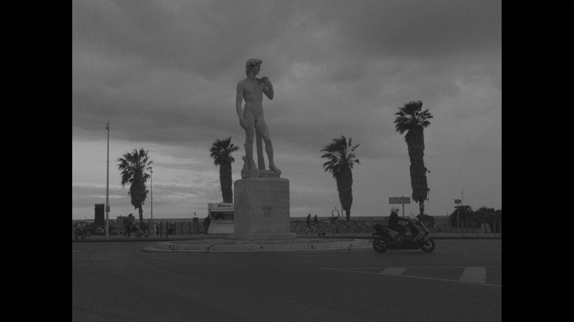 A statue of a male figure standing on a pedestal in a roundabout with palm trees and cloudy sky in the background, with a motorcycle and people visible.