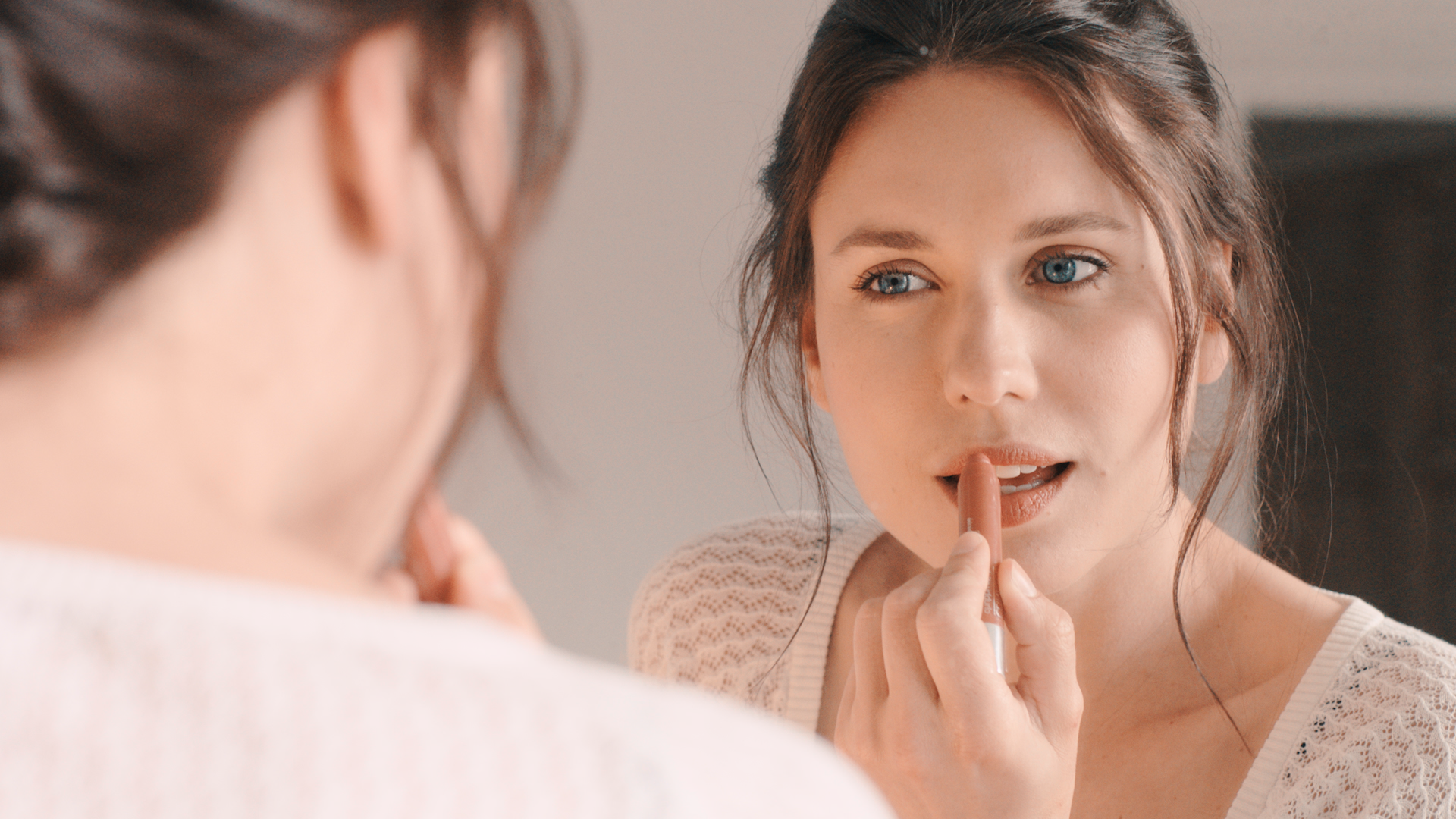 A woman applying nude lipstick while looking into a mirror, with her reflection visible.